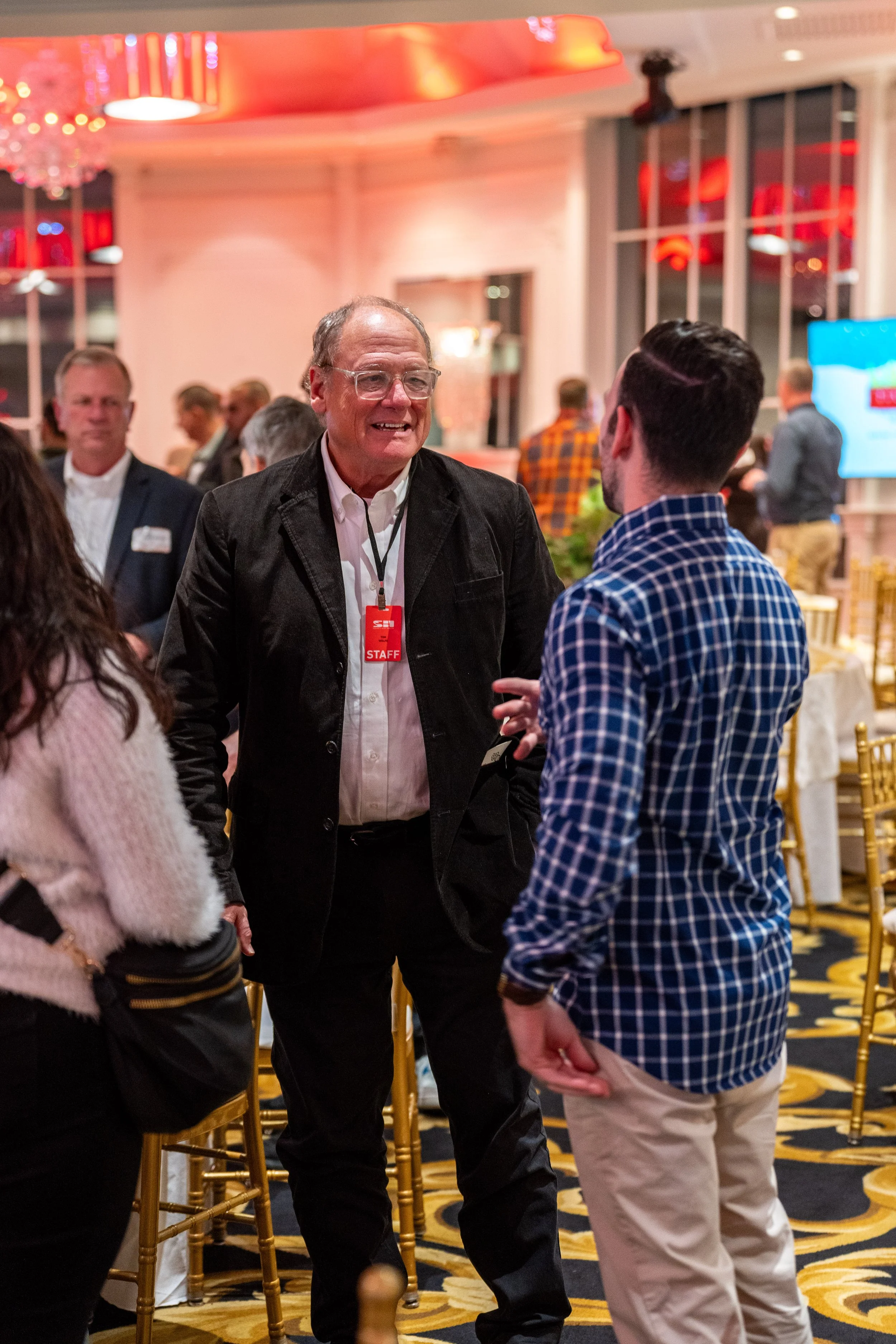People engaged in conversation at a formal event or conference in a decorated banquet room with gold chairs and patterned carpet.