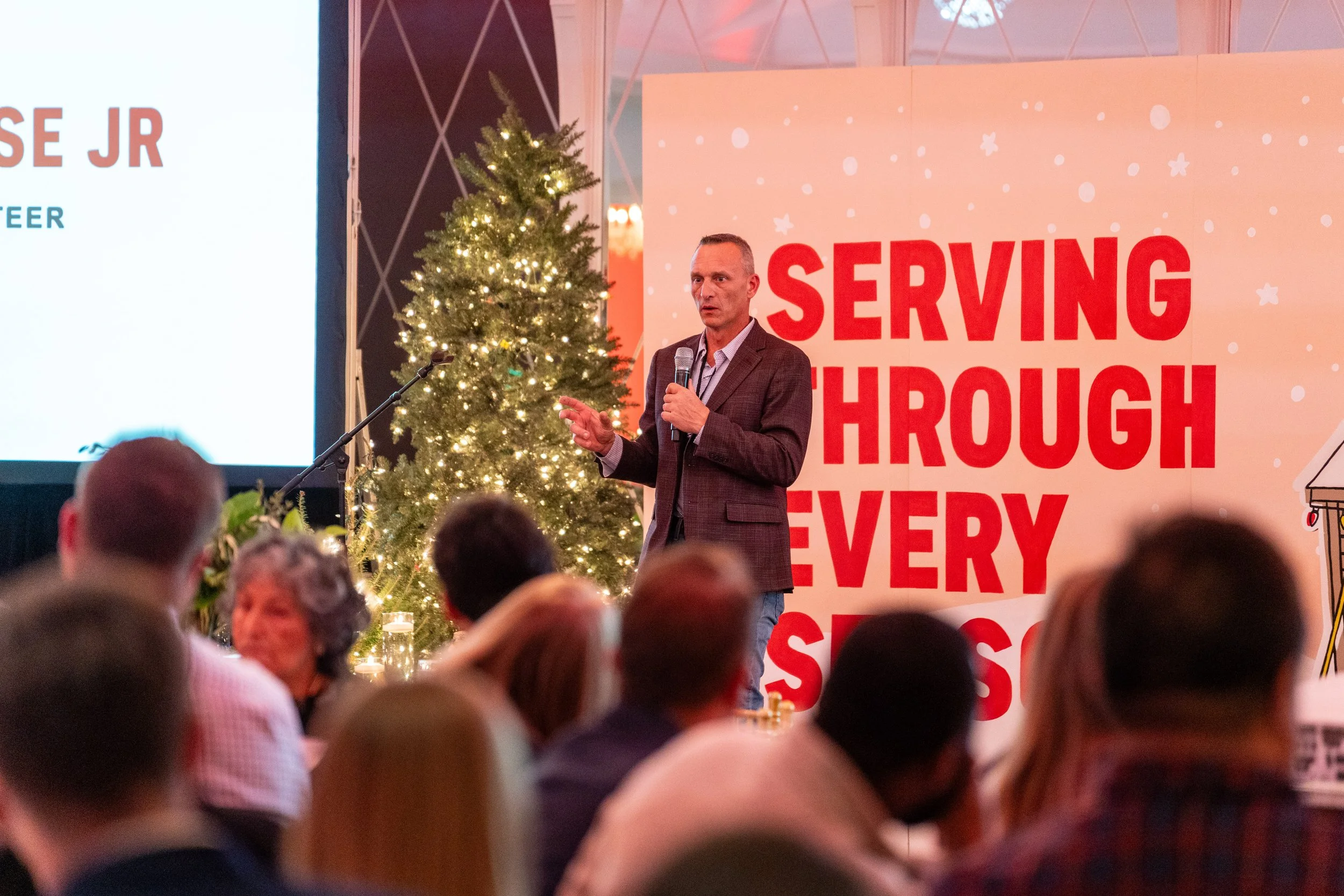Man giving a speech on stage with a Christmas tree and a red and white background reading "Serving through every season."