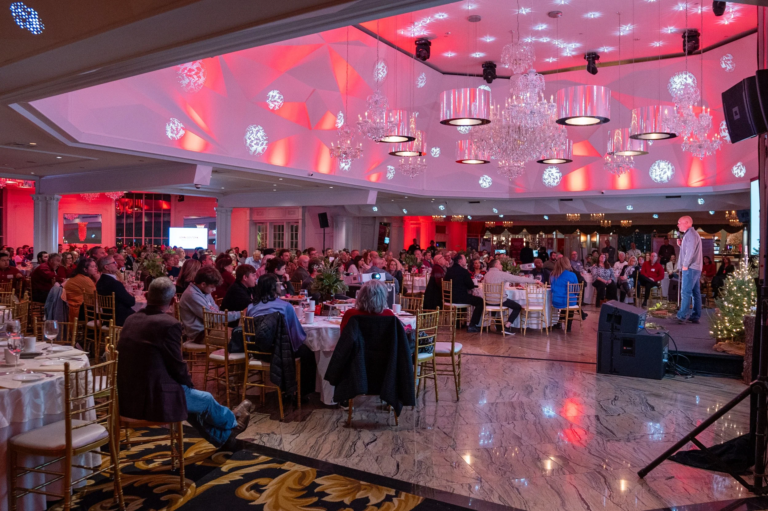 Indoor banquet hall decorated with pink lighting, chandeliers, and snowflake-shaped ceiling lights, filled with seated guests listening to a speaker on stage.