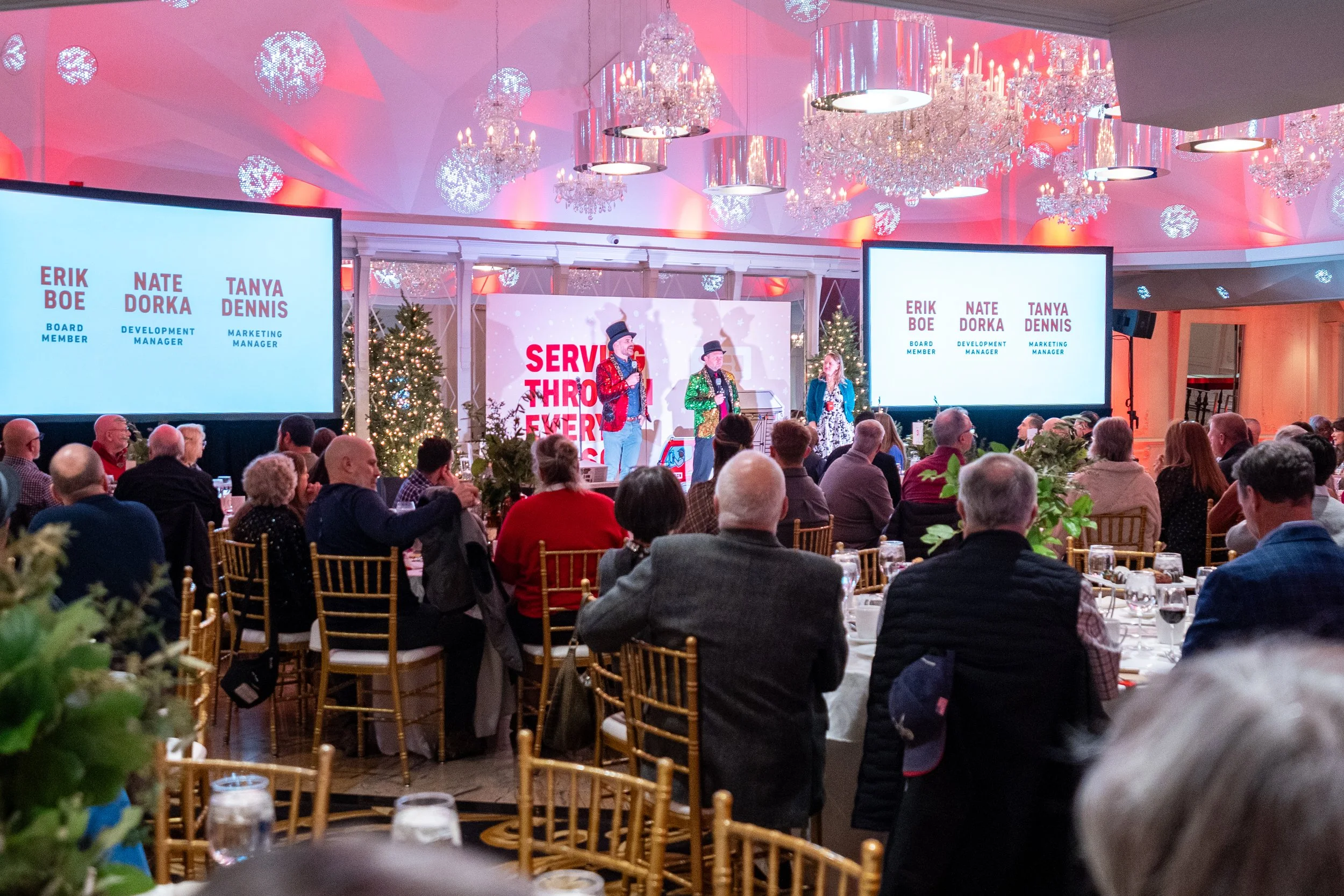 A formal event with people seated at tables in a decorated banquet hall. The stage features three performers in colorful attire, with large screens displaying names and titles of attendees. The ceiling is adorned with chandeliers and holiday lights, 