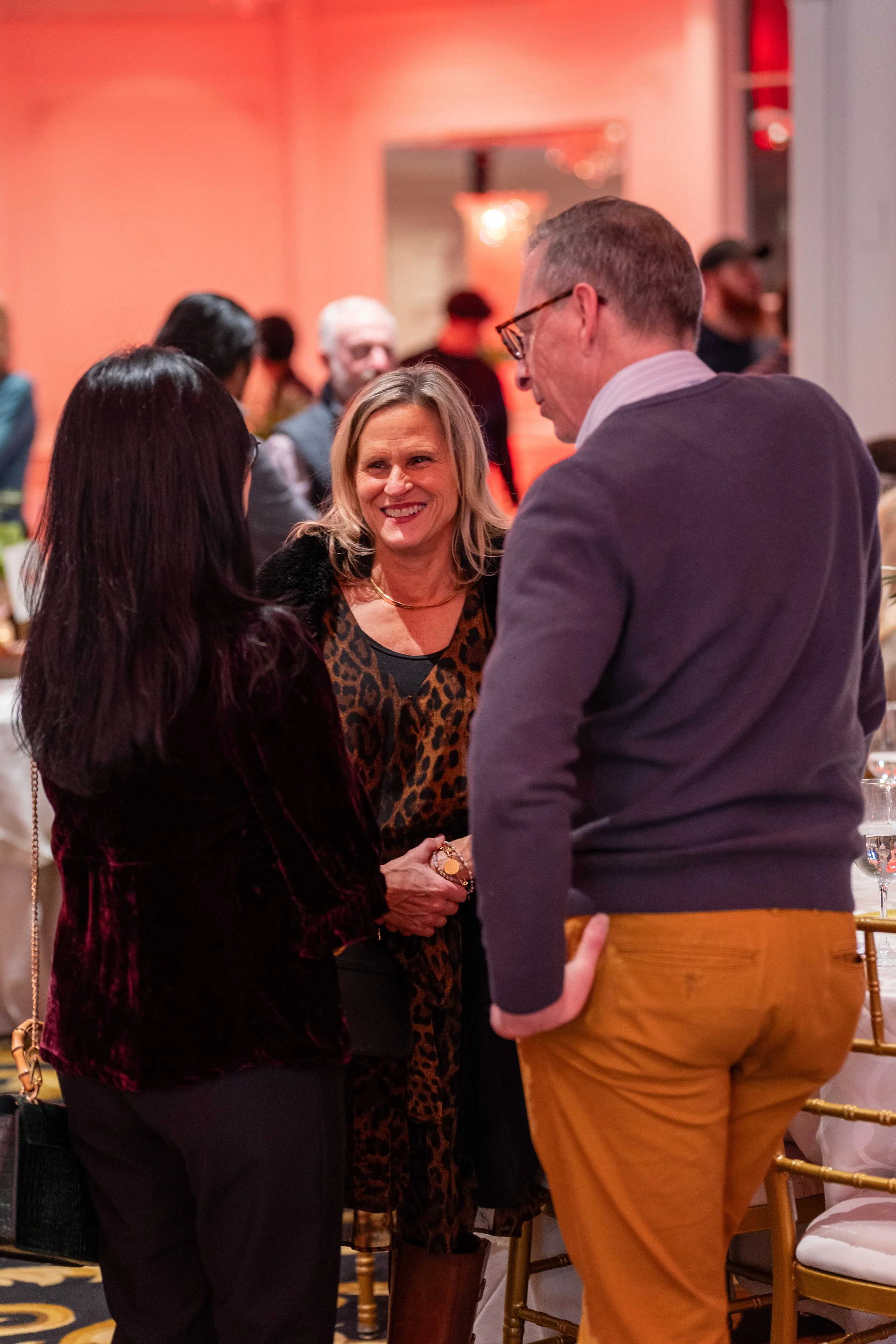 Three people engaged in conversation at a social event with warm orange lighting, with other attendees blurred in the background.