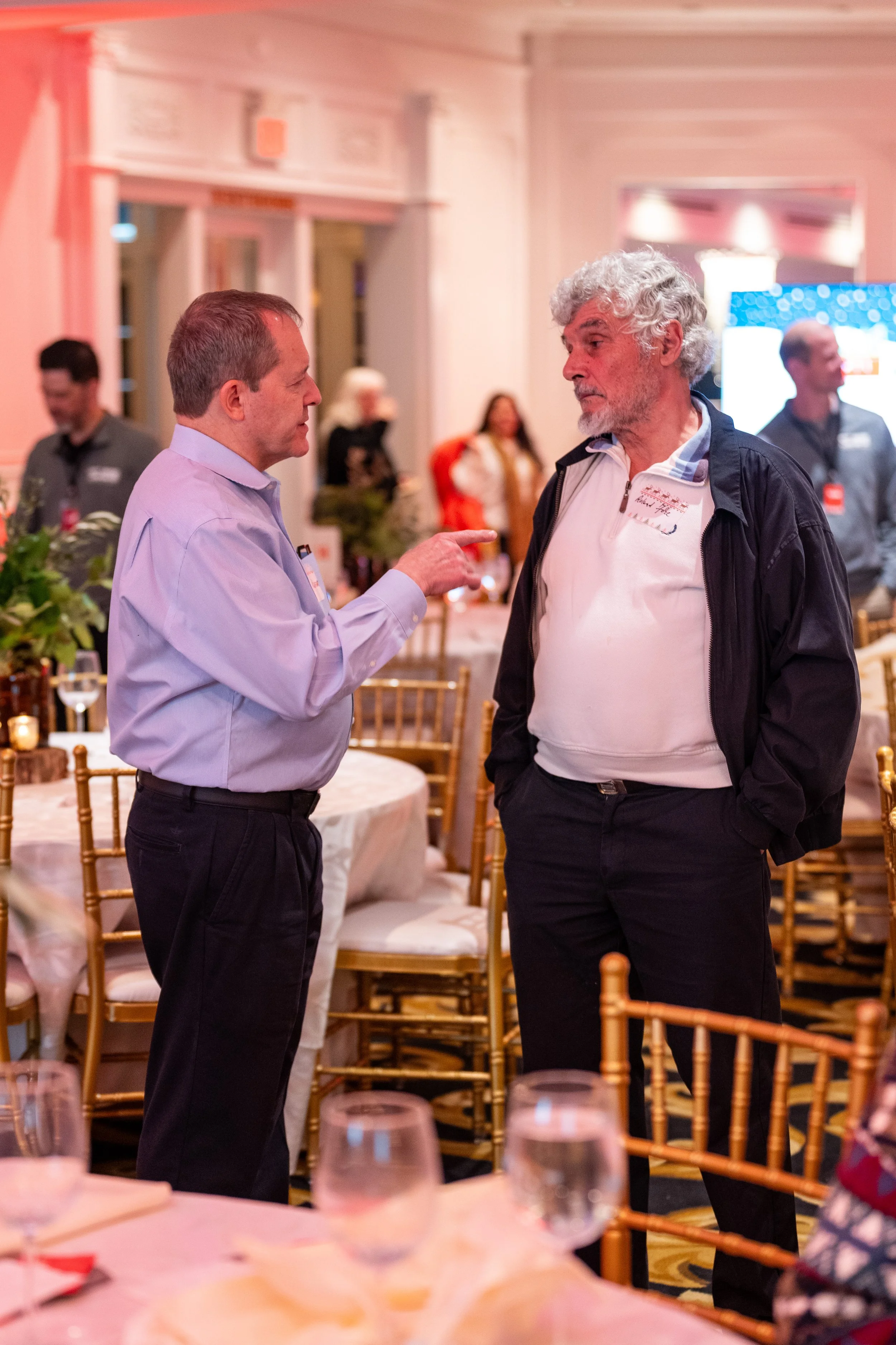 Two men engaged in conversation at a social event, with one gesturing towards the other. The background shows a decorated banquet hall with round tables, chairs, and other attendees.