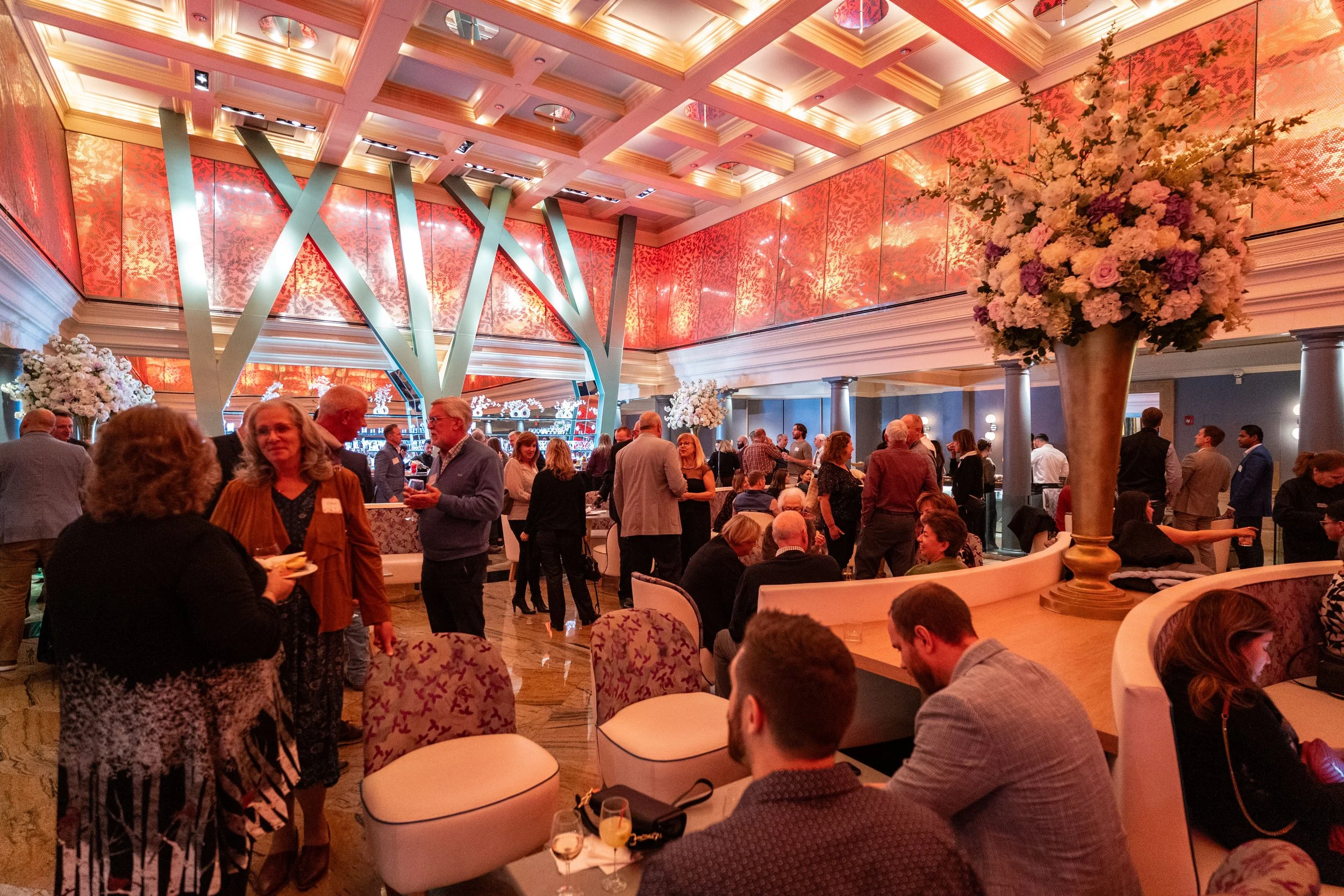 People gathered in a stylish, well-lit event space with floral decorations, modern architecture, and a bar area in the background.
