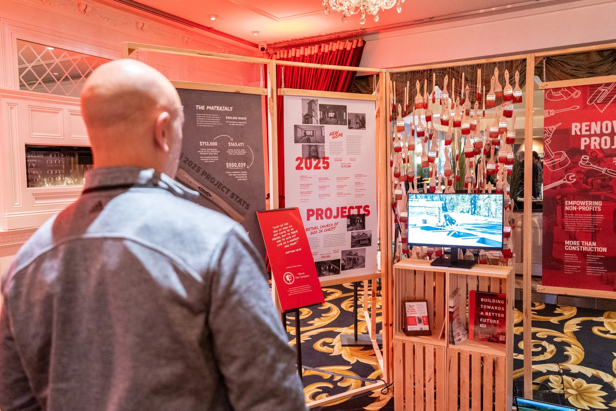 A man observes a display booth with project information, charts, and images at an indoor event space with carpeted floors and red curtains.