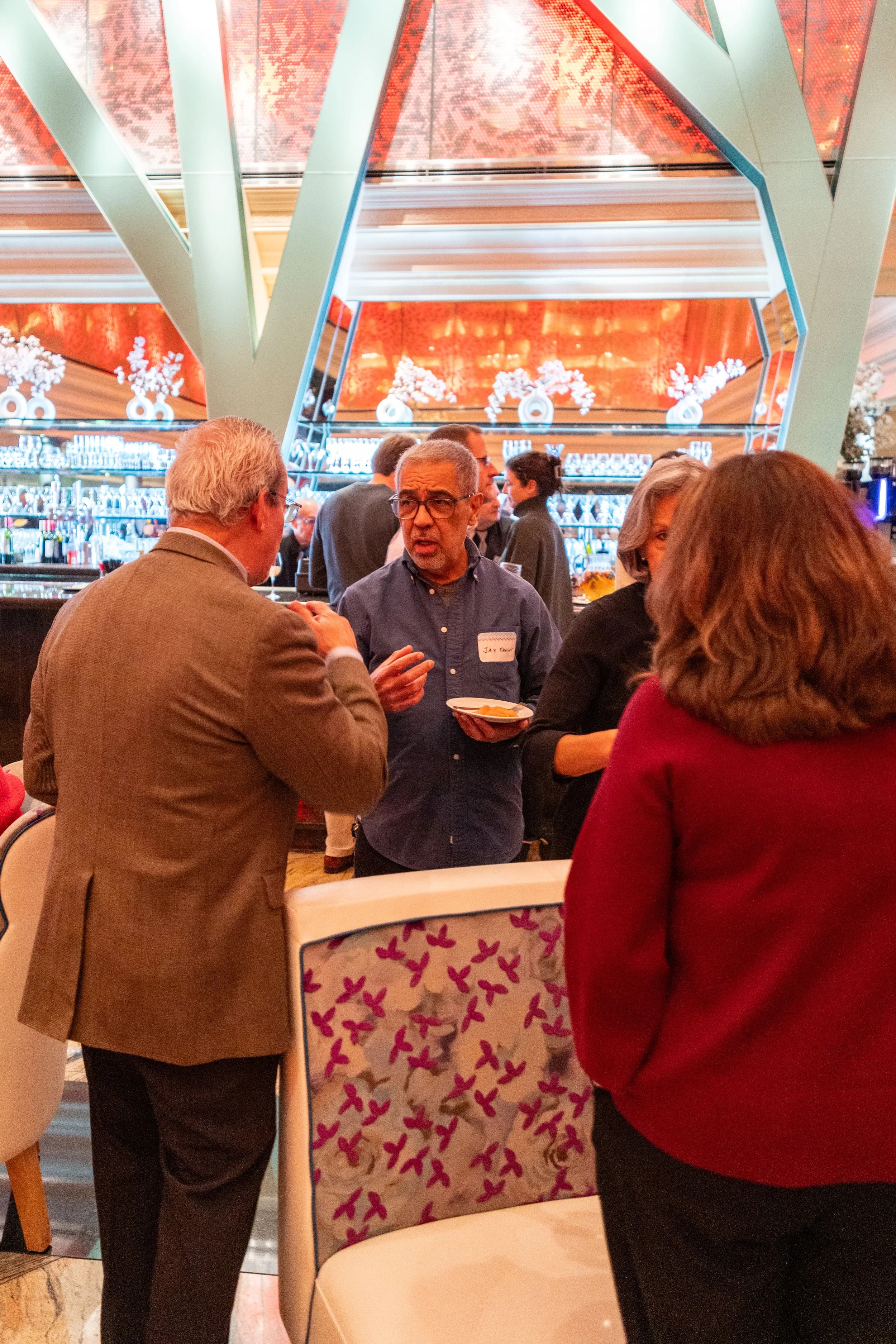 People engaging in conversations at a bar or social event in an elegant venue, with a colorful and modern interior decor including floral arrangements and high ceilings.