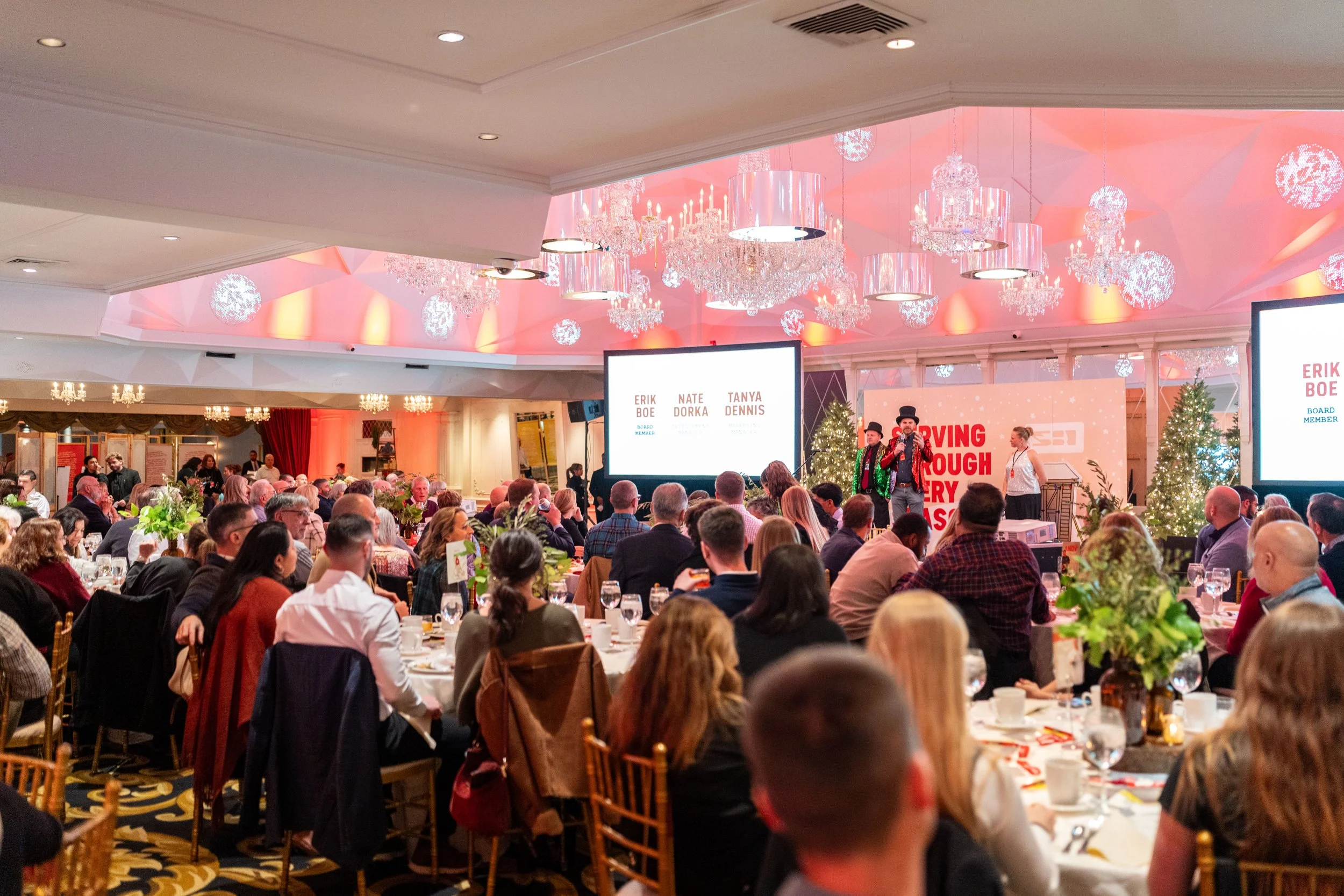 A large banquet hall filled with seated attendees at round tables, with a stage at the front where performers dressed in black and red are entertaining. The room is decorated with Christmas trees, chandeliers, and festive lighting, and large screens 