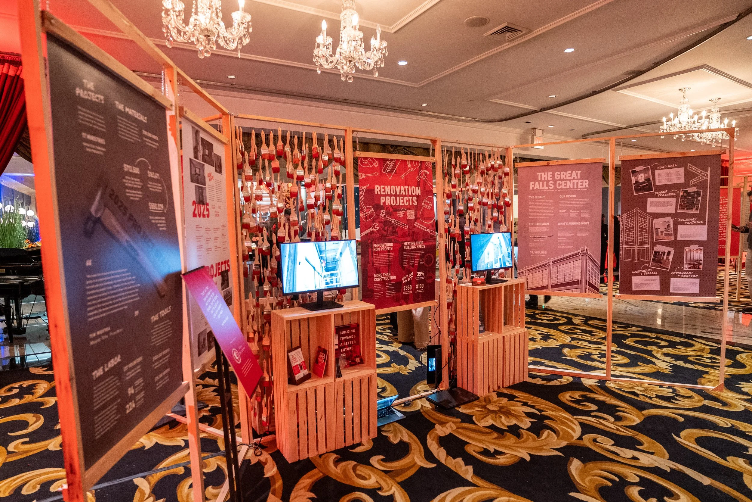 An exhibition setup with wooden frame panels displaying informational posters and two computer monitors. The posters discuss renovation projects, the Great Falls Center, and the culture of the building. The event is indoors with a richly patterned ca