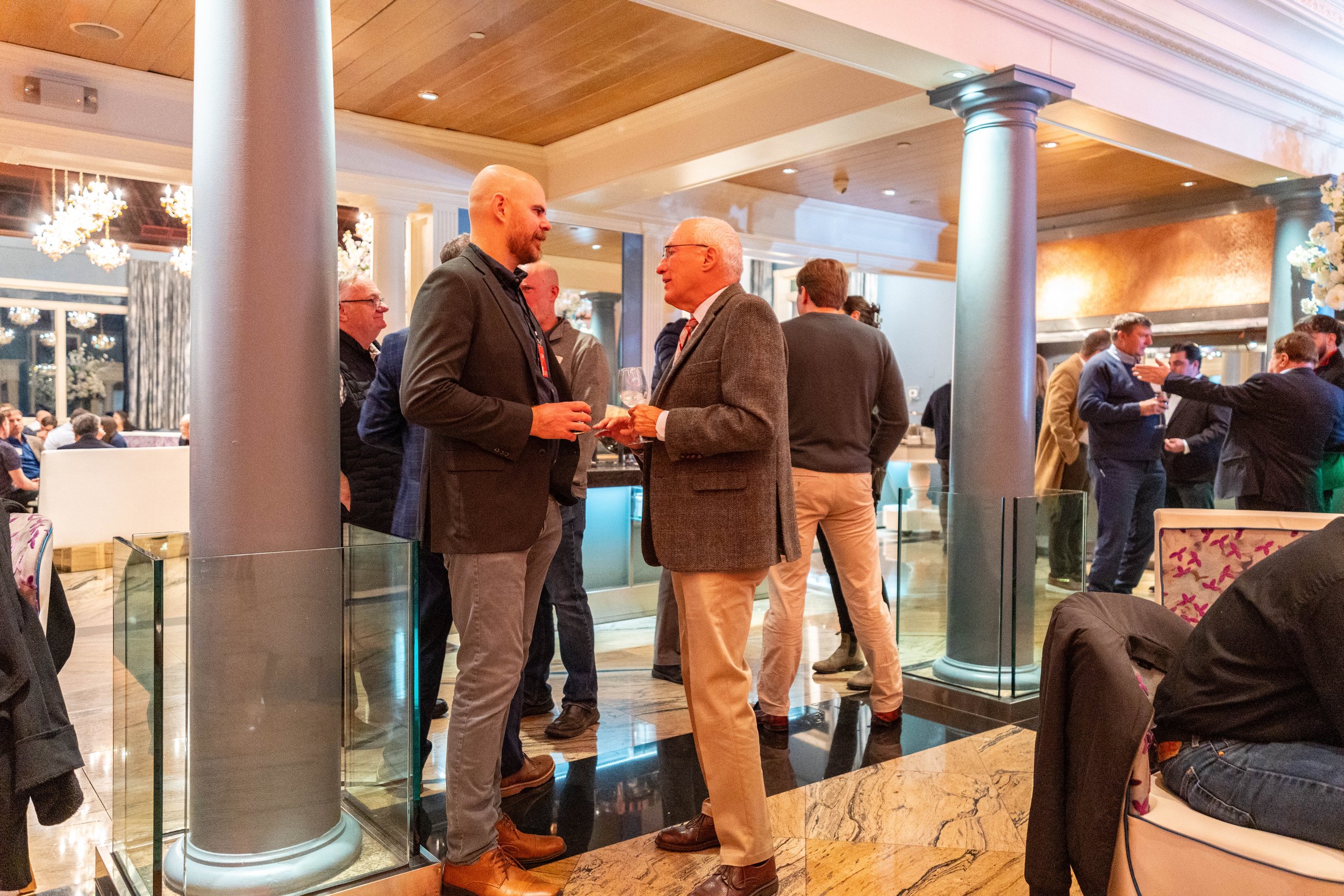 Two men in business attire conversing at a social event, holding drinks, in a elegantly decorated indoor venue with other attendees in the background.