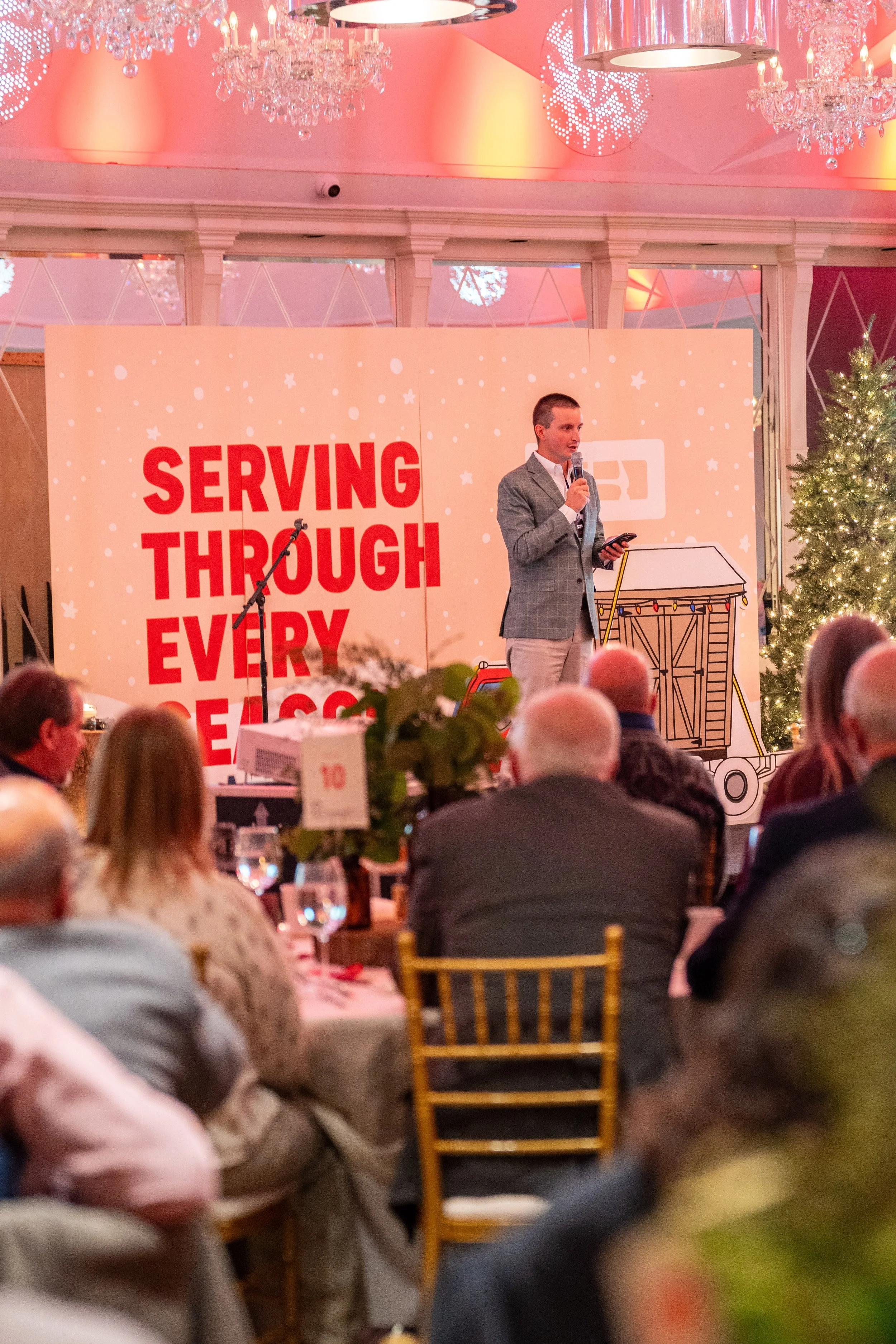 A man in a gray checkered blazer speaks into a microphone on stage at a formal event, with a Christmas tree nearby, and a backdrop that reads 'Serving Through Every Season.' The audience is seated at round tables decorated with candles and table numb