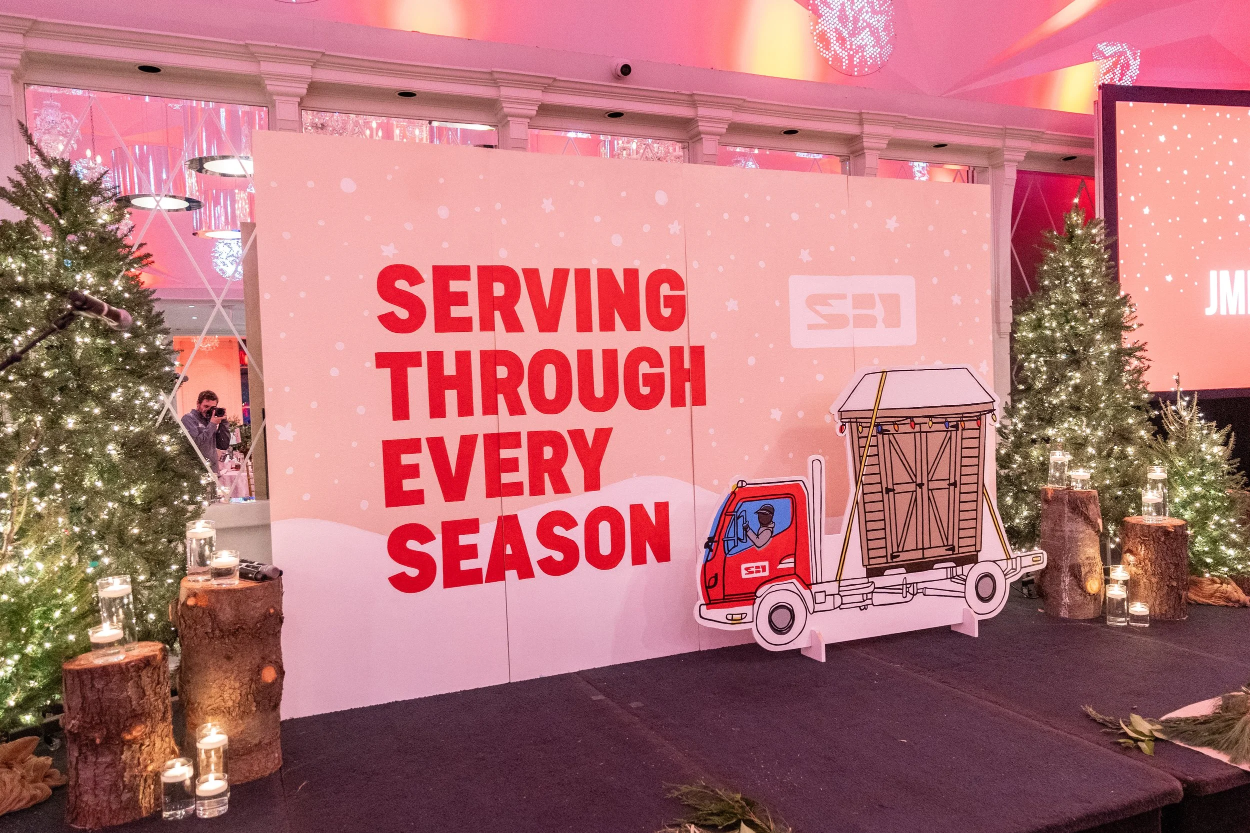 A holiday-themed display with a large sign reading "Serving Through Every Season" in red letters, decorated with Christmas trees, candles on logs, and a cardboard cutout of a truck with a shed on it.