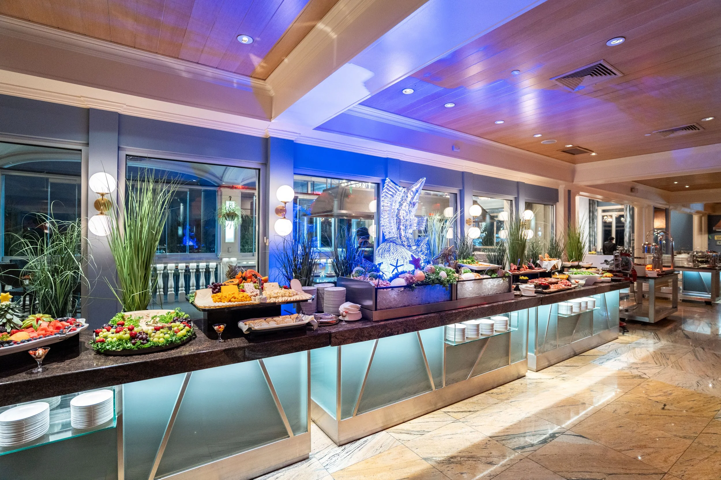Buffet table with various food dishes, decorated with plants and a large illuminated underwater scene with a fish sculpture in the center, inside a well-lit dining area.