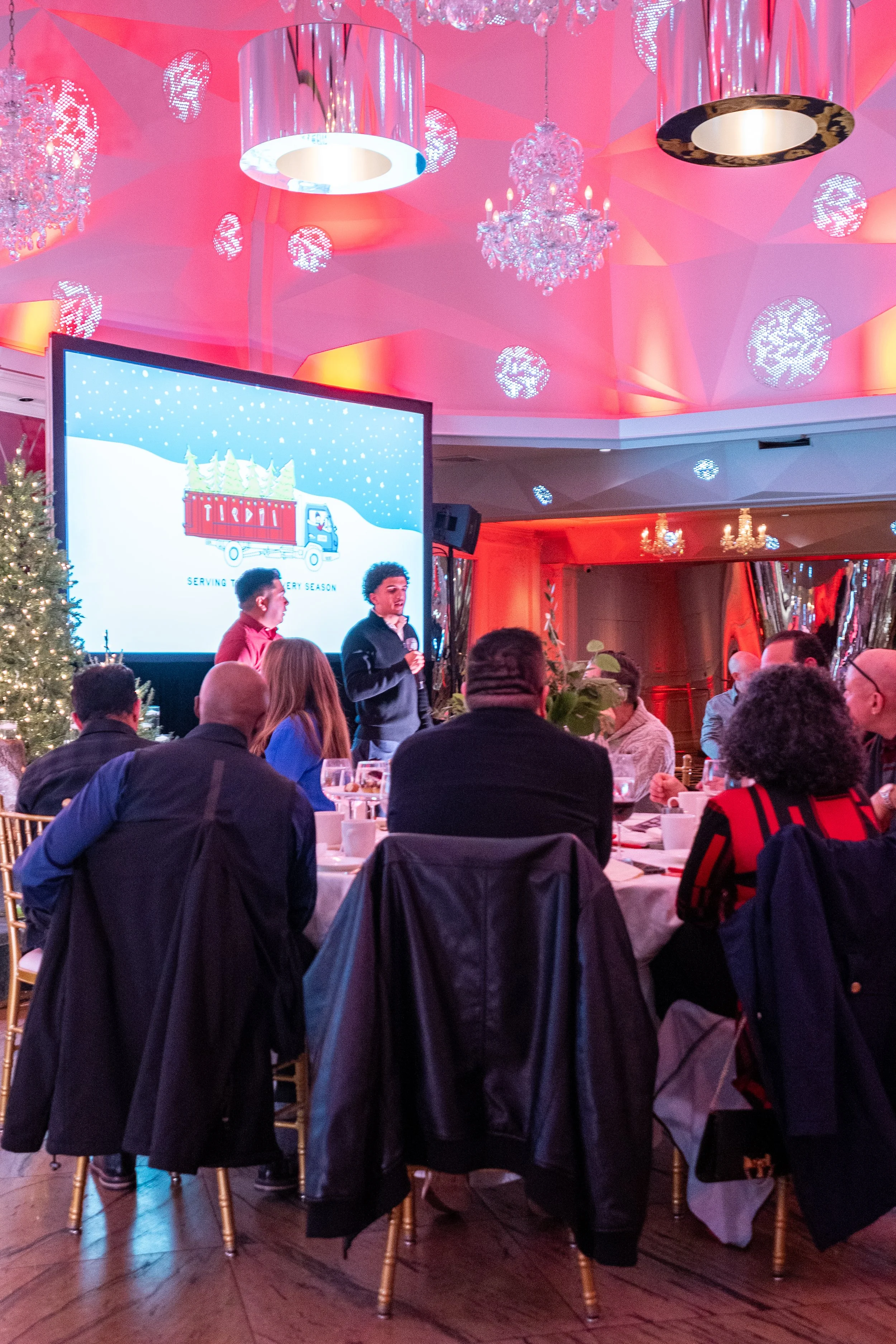 People sitting at round tables during a holiday event, with a presenter speaking in front of a large screen displaying a holiday graphic, in a decorated venue with Christmas trees, chandeliers, and colorful lighting.