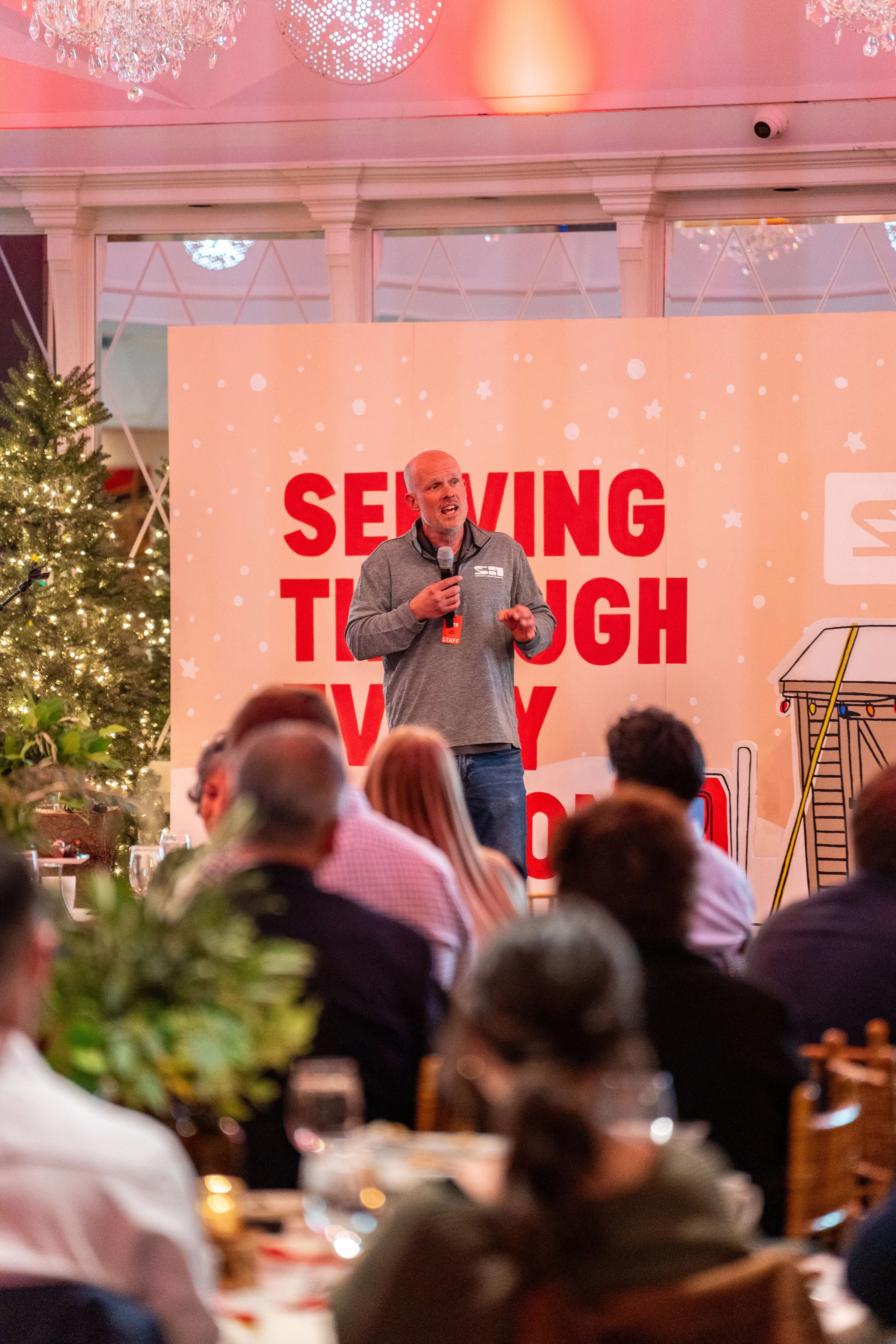 A man speaking into a microphone at a conference with a backdrop that reads 'SERVING THROUGH THE V' and festive holiday decorations, including a Christmas tree with lights, in a decorated indoor venue.