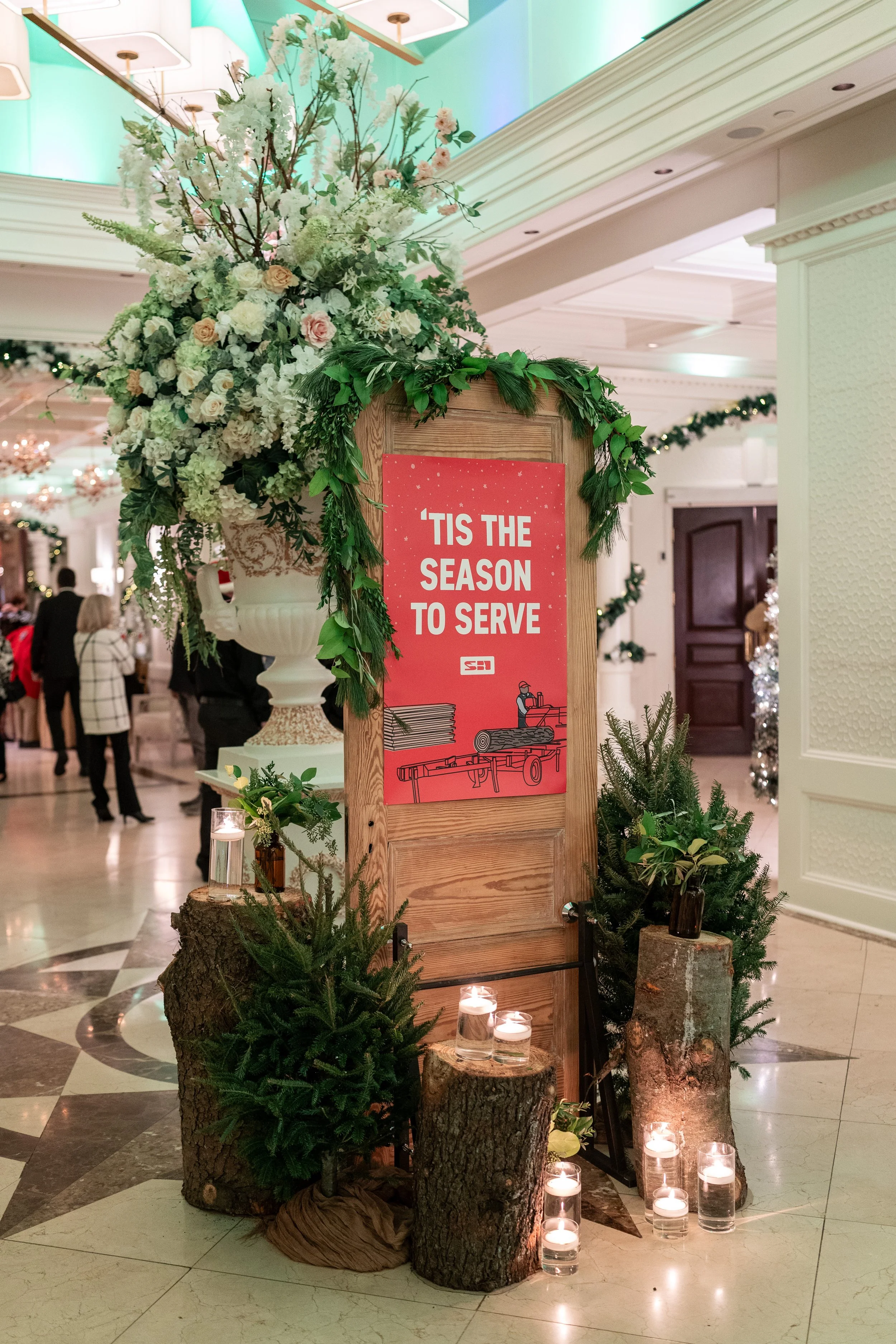A Christmas-themed sign with the text 'Tis the season to serve' on a wooden stand, decorated with green foliage, small Christmas trees, candles, and a large floral arrangement in a white vase, set in an elegant indoor venue with holiday decorations a