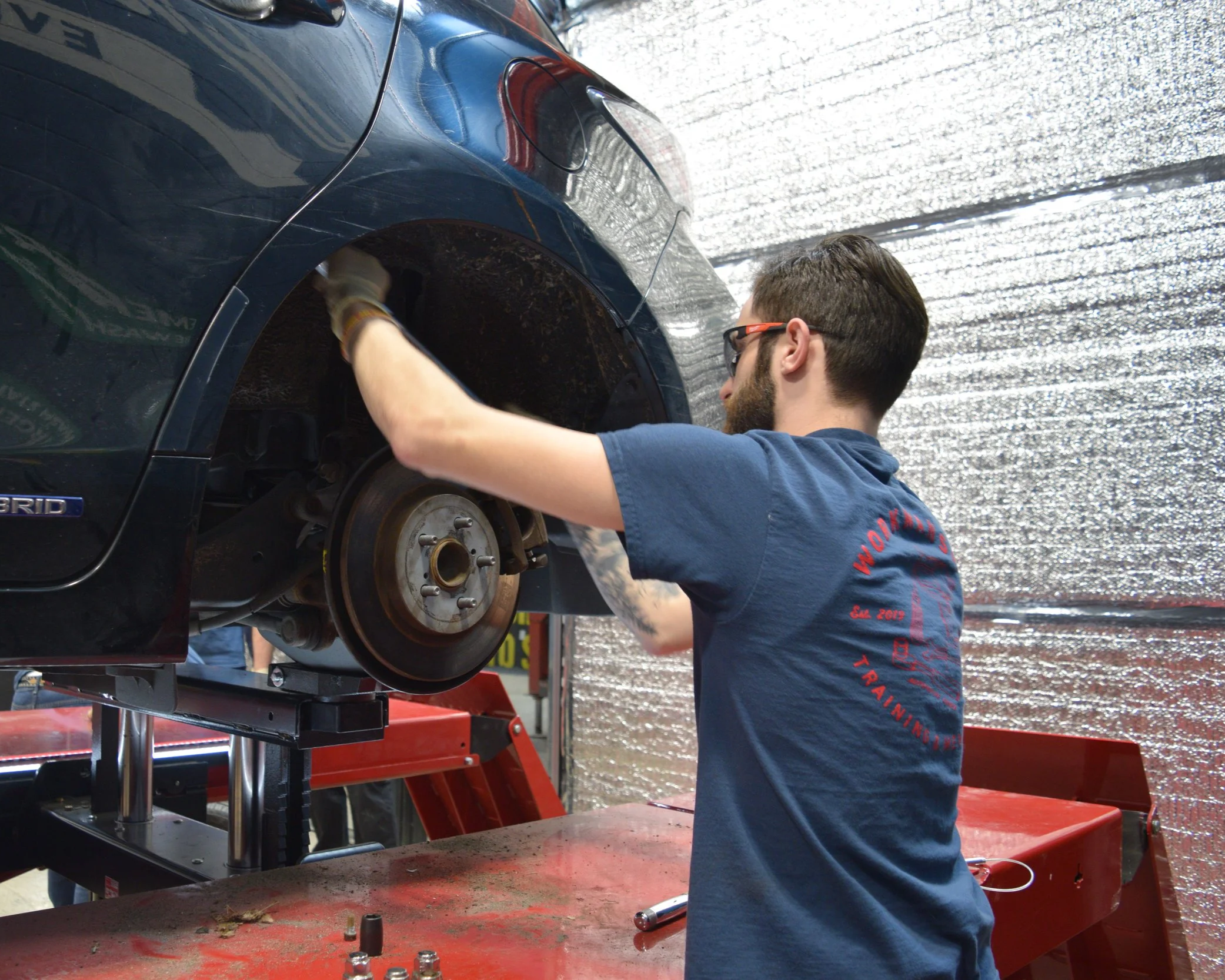 A man with glasses and a beard working on the brake system of a blue hybrid car, elevated on a lift in an auto repair shop.