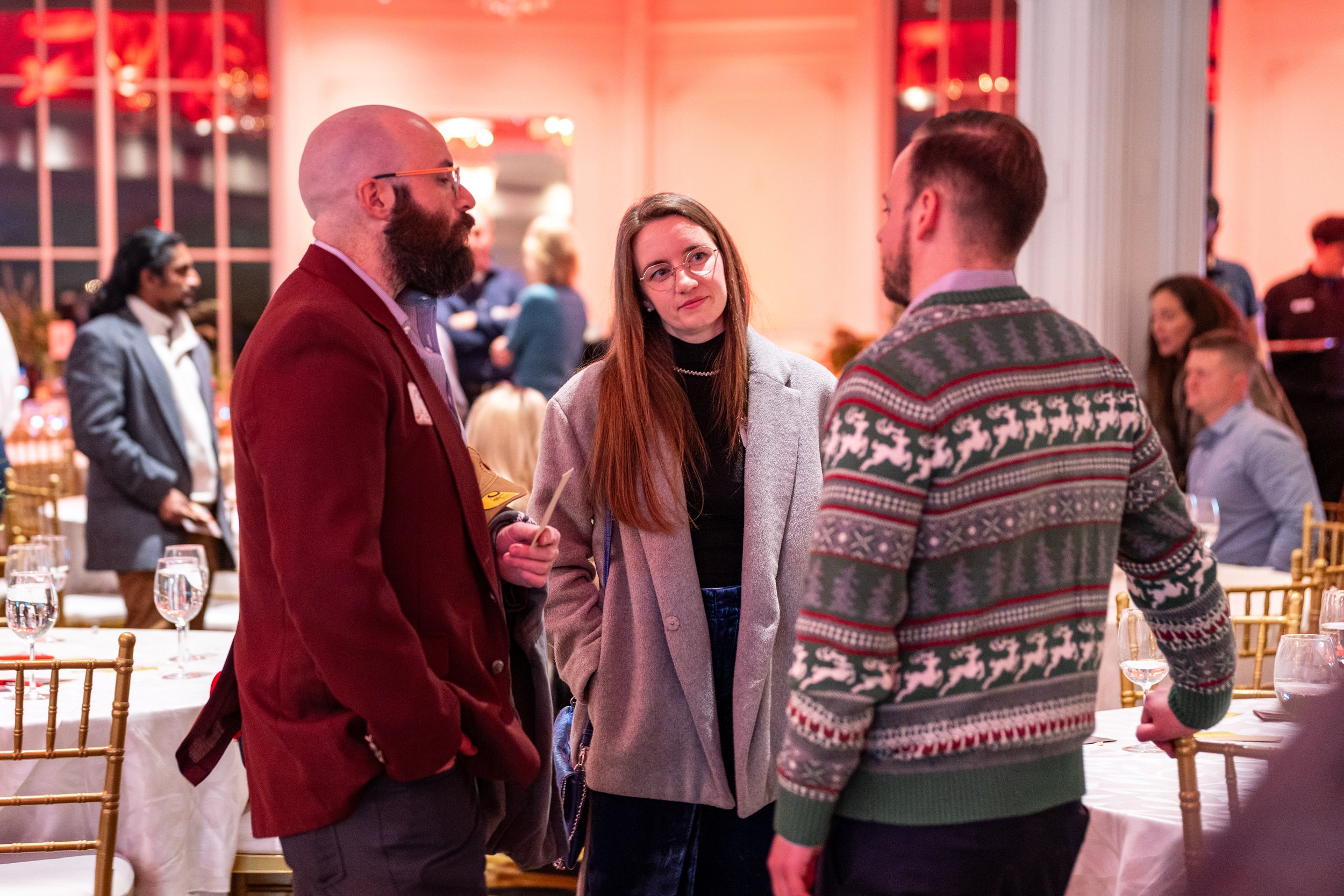 Three people in formal attire engaged in conversation at a festive event, with tables, chairs, and other attendees in the background.