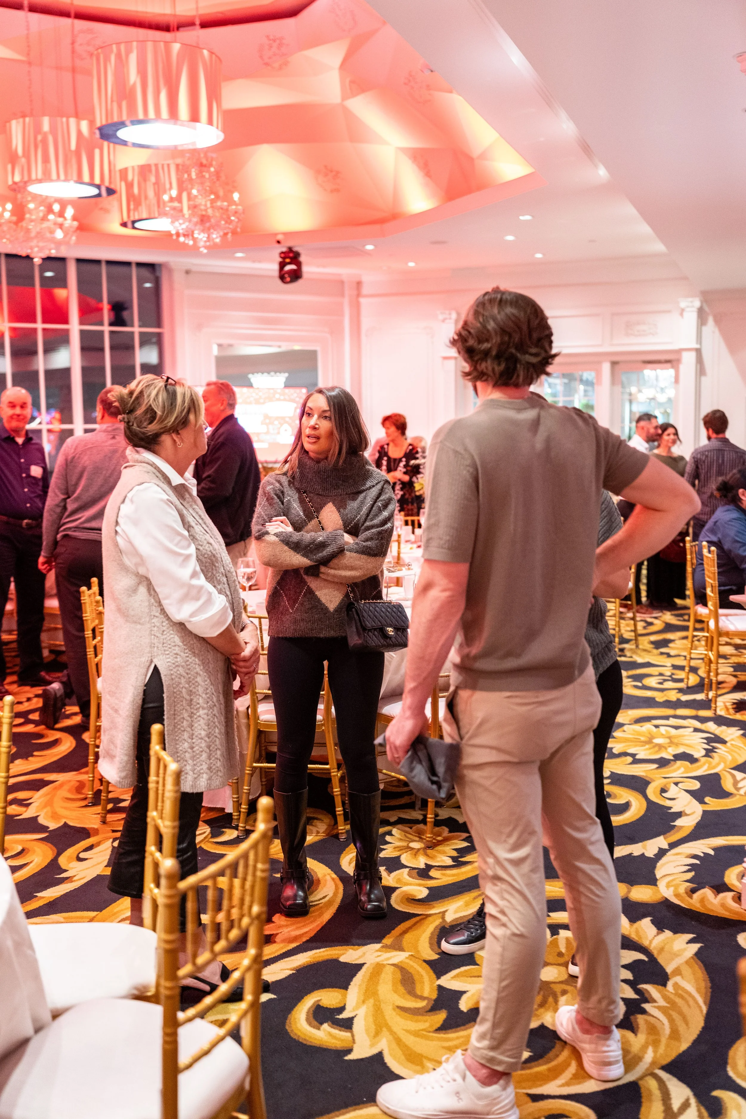 Group of people chatting at a formal event in an elegantly decorated ballroom with gold chairs, patterned carpet, and pink lighting.
