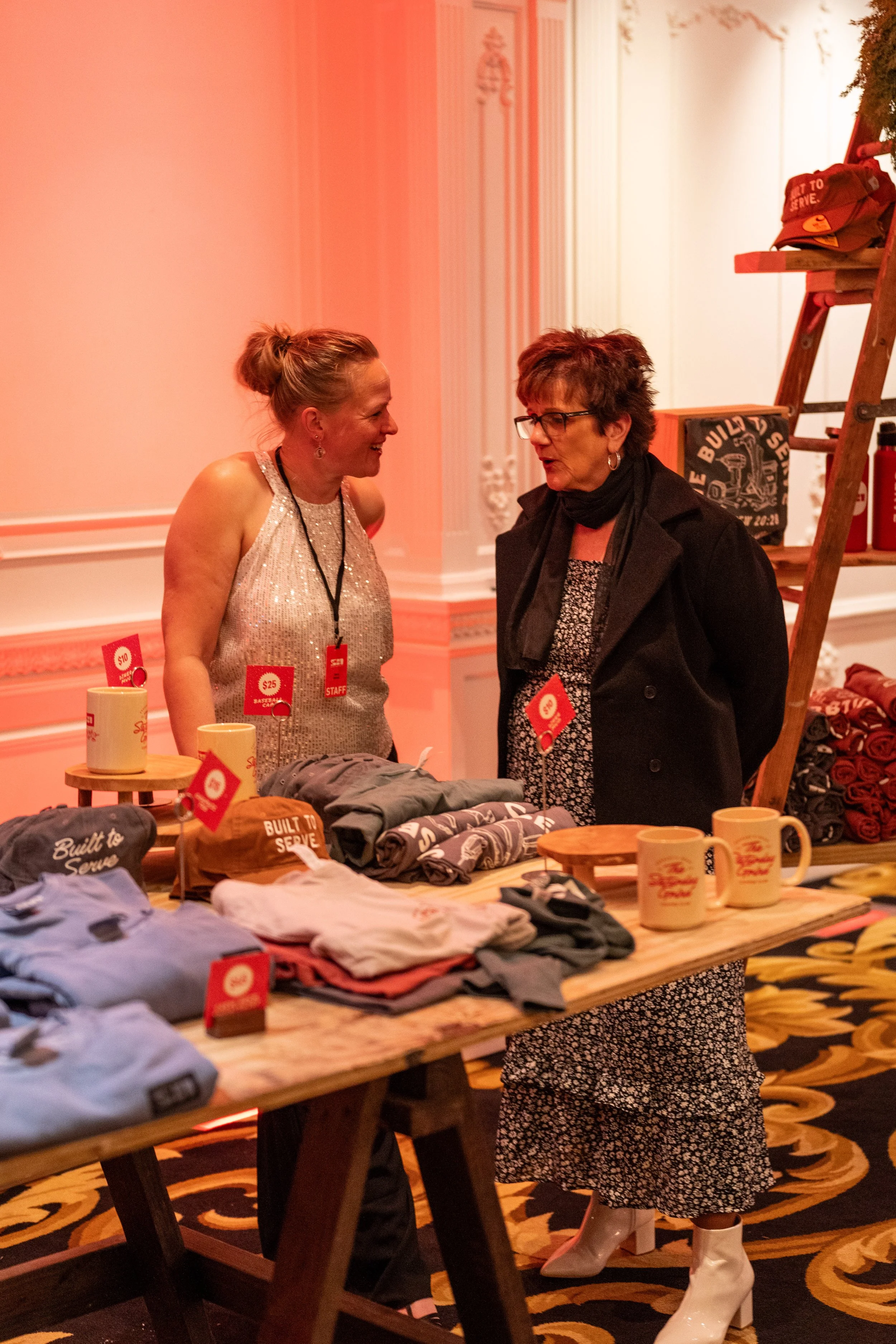Two women talking at a merchandise booth with sweatshirts, mugs, and hats displayed on a wooden table and shelf.