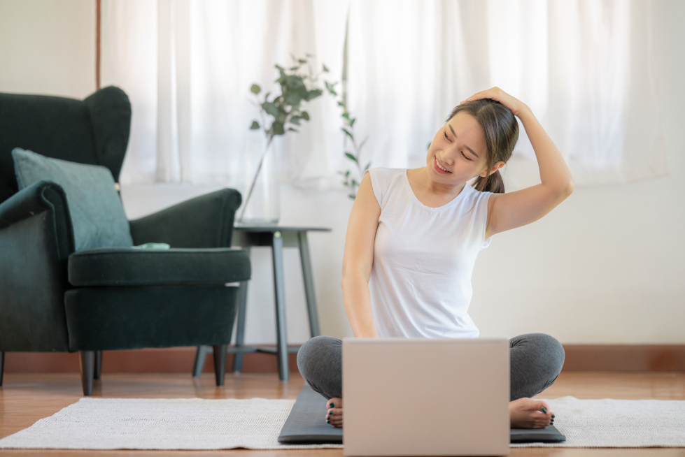 Woman sitting on a yoga mat in front of a laptop doing a neck stretch