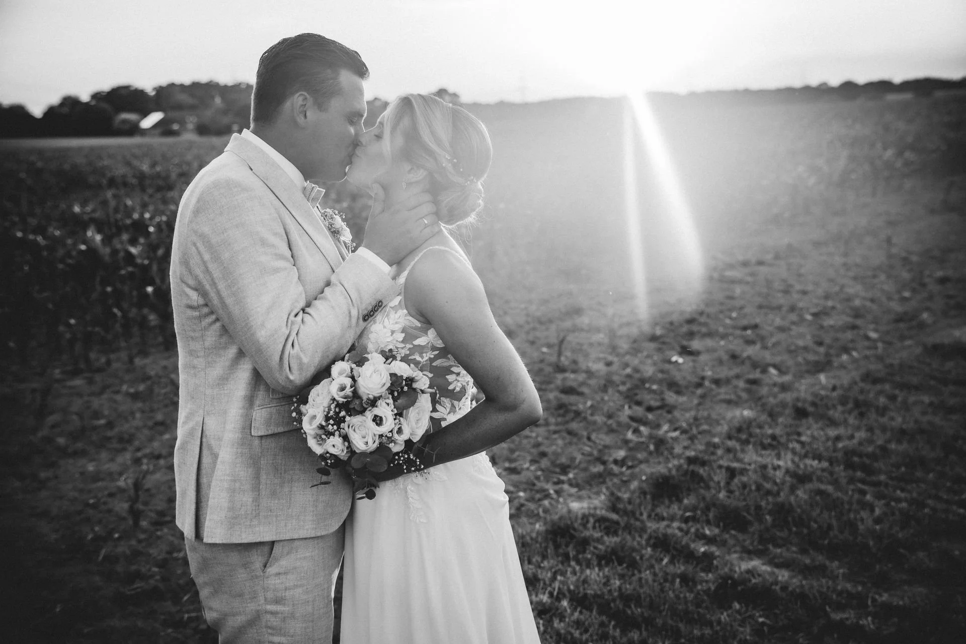 Ein Hochzeitsbild in Schwarz-Weiß von einem Paar, das sich küssend im Freien bei Sonnenuntergang befindet, mit einer Frau in einem Hochzeitskleid und einem Mann in einem Anzug, beide liebevoll verbunden.