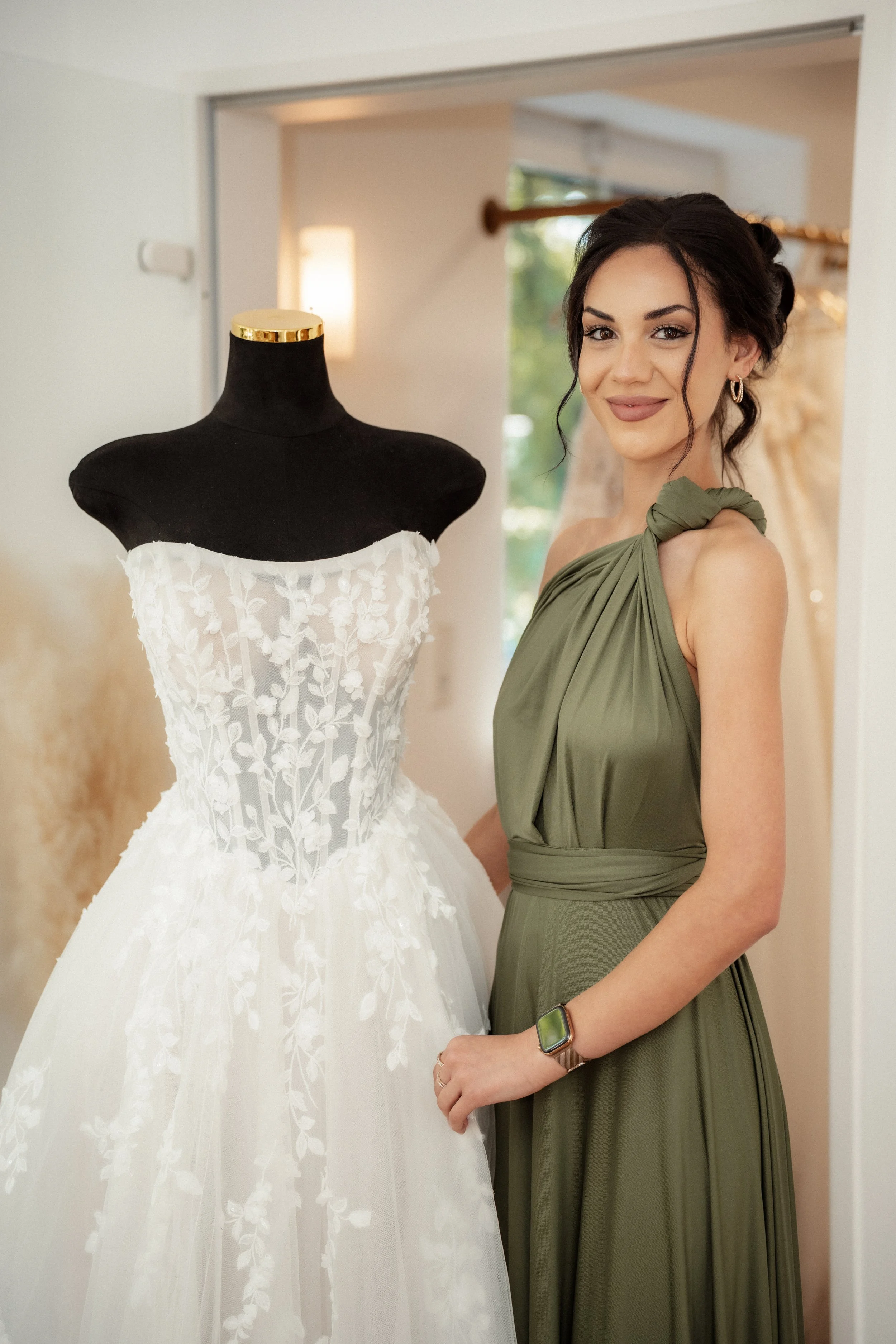 Eine junge Frau in einem grünem Kleid steht neben einem Brautkleid auf einem schwarzen Modellständer, in einem gut beleuchteten Raum mit natürlichem Licht.