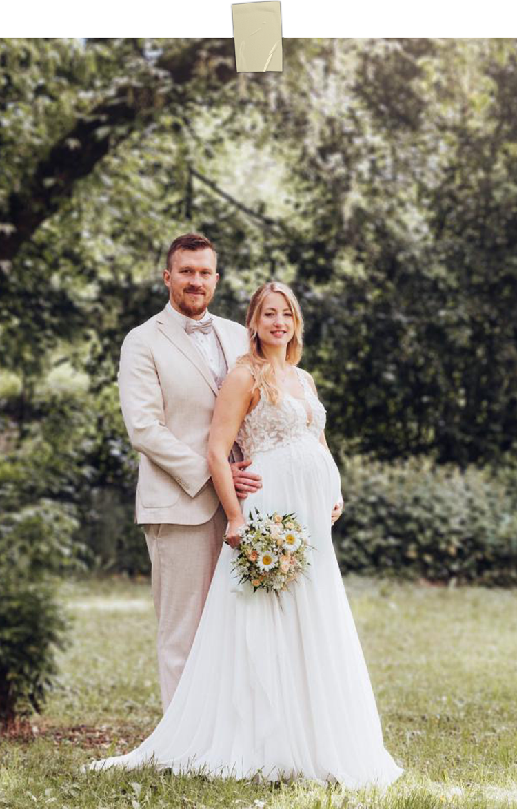 Ein Brautpaar steht im Park, die Frau trägt ein weißes Brautkleid und hält einen Blumenstrauß, der Mann trägt einen hellen Anzug. Im Hintergrund sind Bäume und grüne Vegetation.