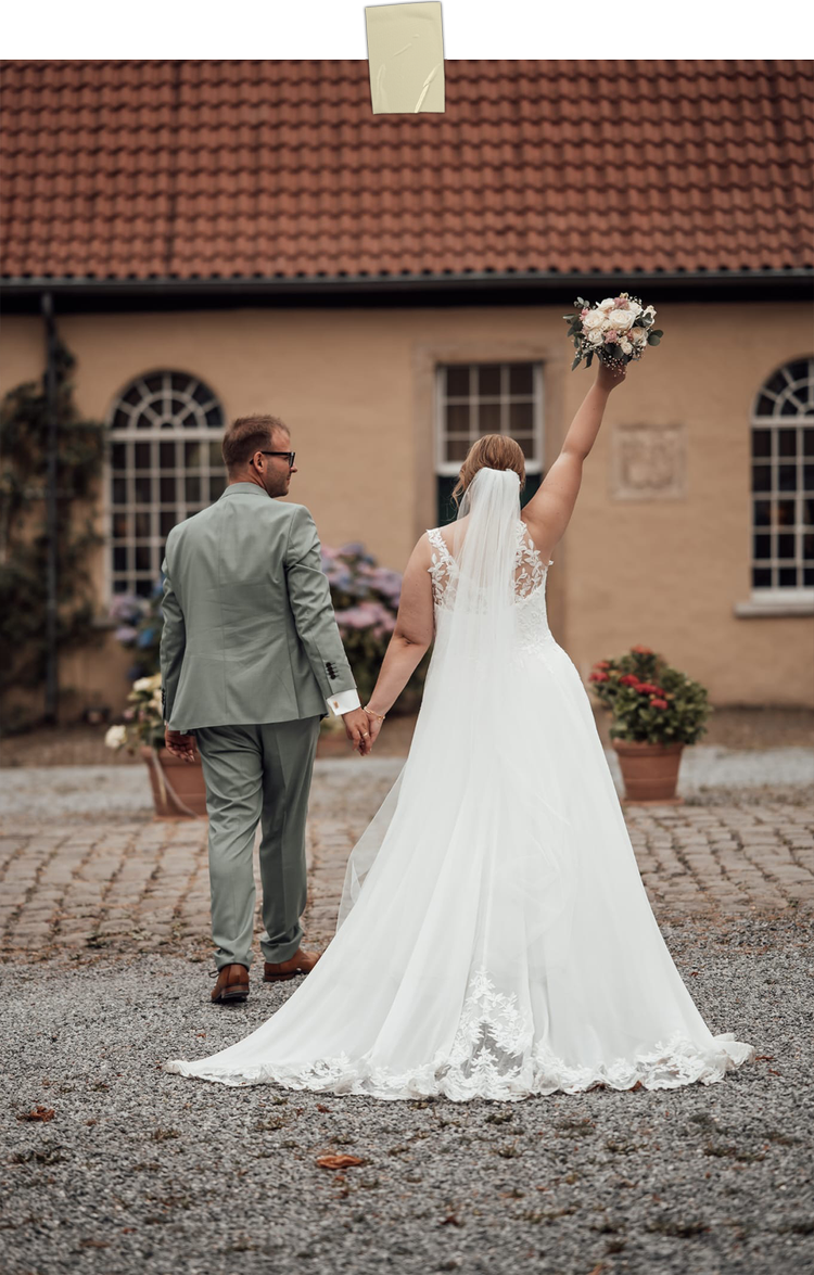 Ein Brautpaar beim Hochzeitsspaziergang vor einem alten Haus mit roten Ziegeln, die Braut trägt ein weißes Brautkleid mit Spitzendetails und hält einen Blumenstrauß hoch, während sie die Hand des Bräutigams hält.