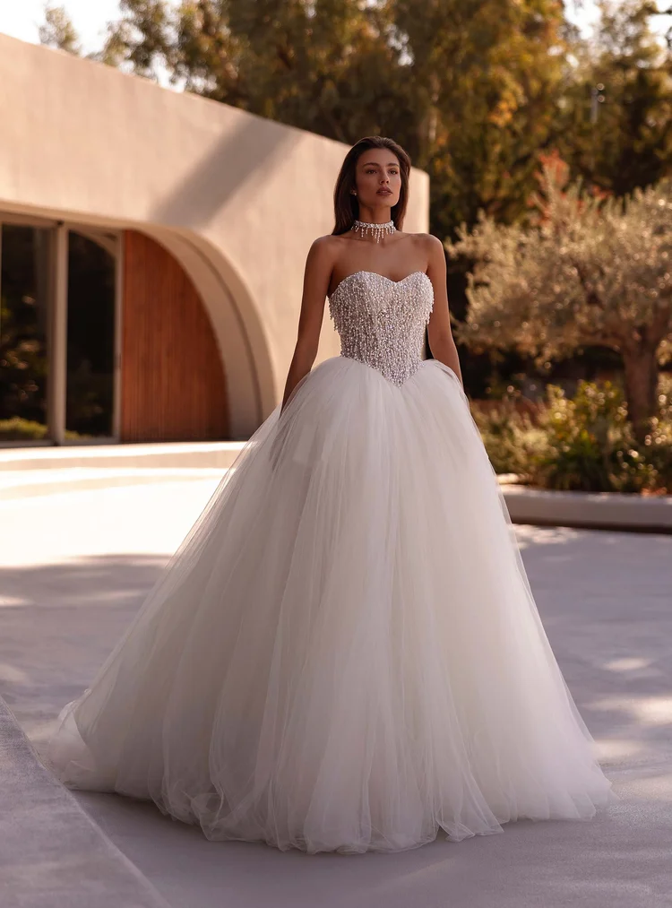 Eine Frau in einem Brautkleid mit Schleier, Schmuck und einer hohen Tüllrock steht vor einer modernen, weißen Architektur im Freien bei Sonnenuntergang.