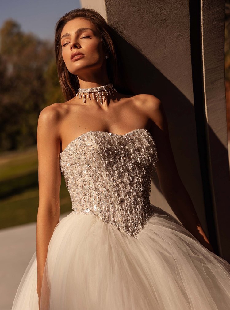 Frau in einem eleganten, perlenverzierten Brautkleid mit einem Tüllrock, der an einem sonnigen Tag im Freien steht und die Augen geschlossen hat.