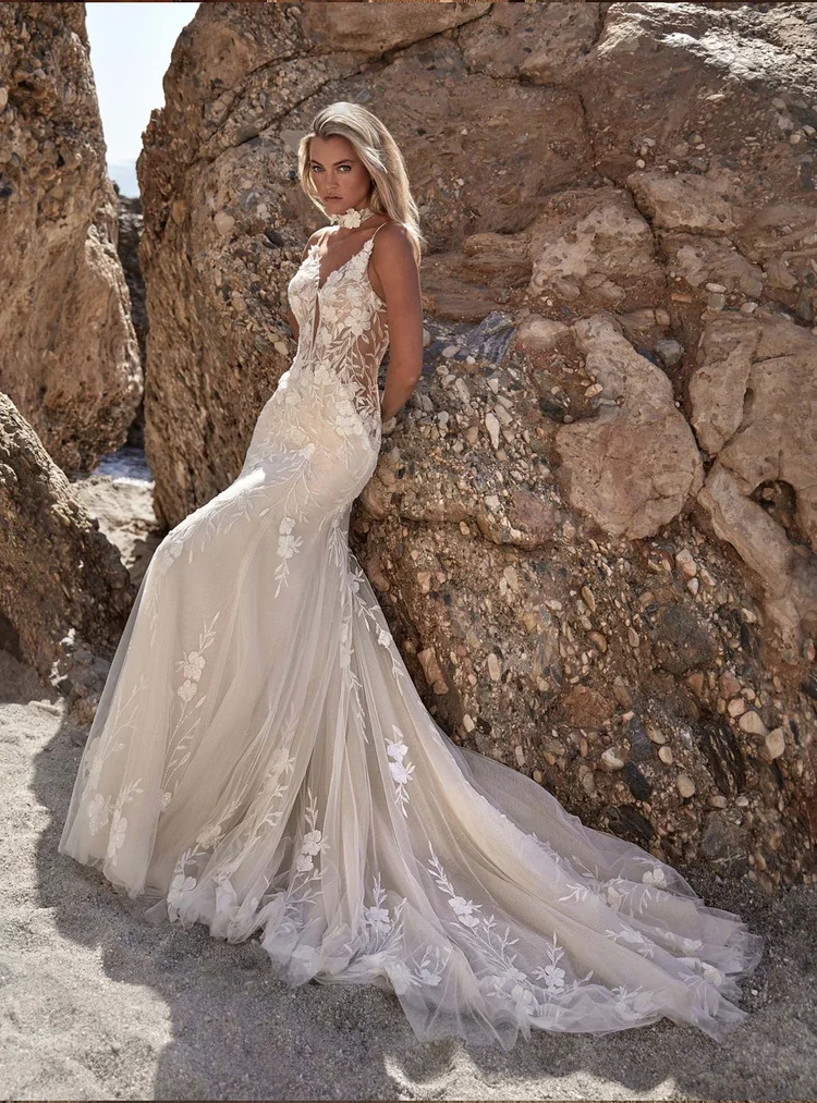 Eine Frau in einem weißen Hochzeitskleid mit Stickerei steht an einem Strand zwischen Felsen.