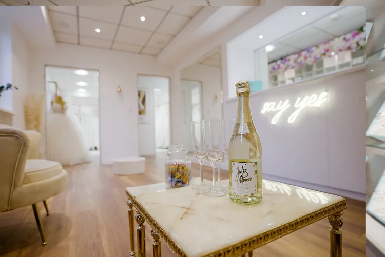 Ein mit einem goldfarbenen Rahmen verzierter Tisch mit Sektflasche, Gläsern und Süßigkeiten in einem hellen, modern eingerichteten Raum. Im Hintergrund ein beleuchteter Schriftzug 'say yes' an der Wand.