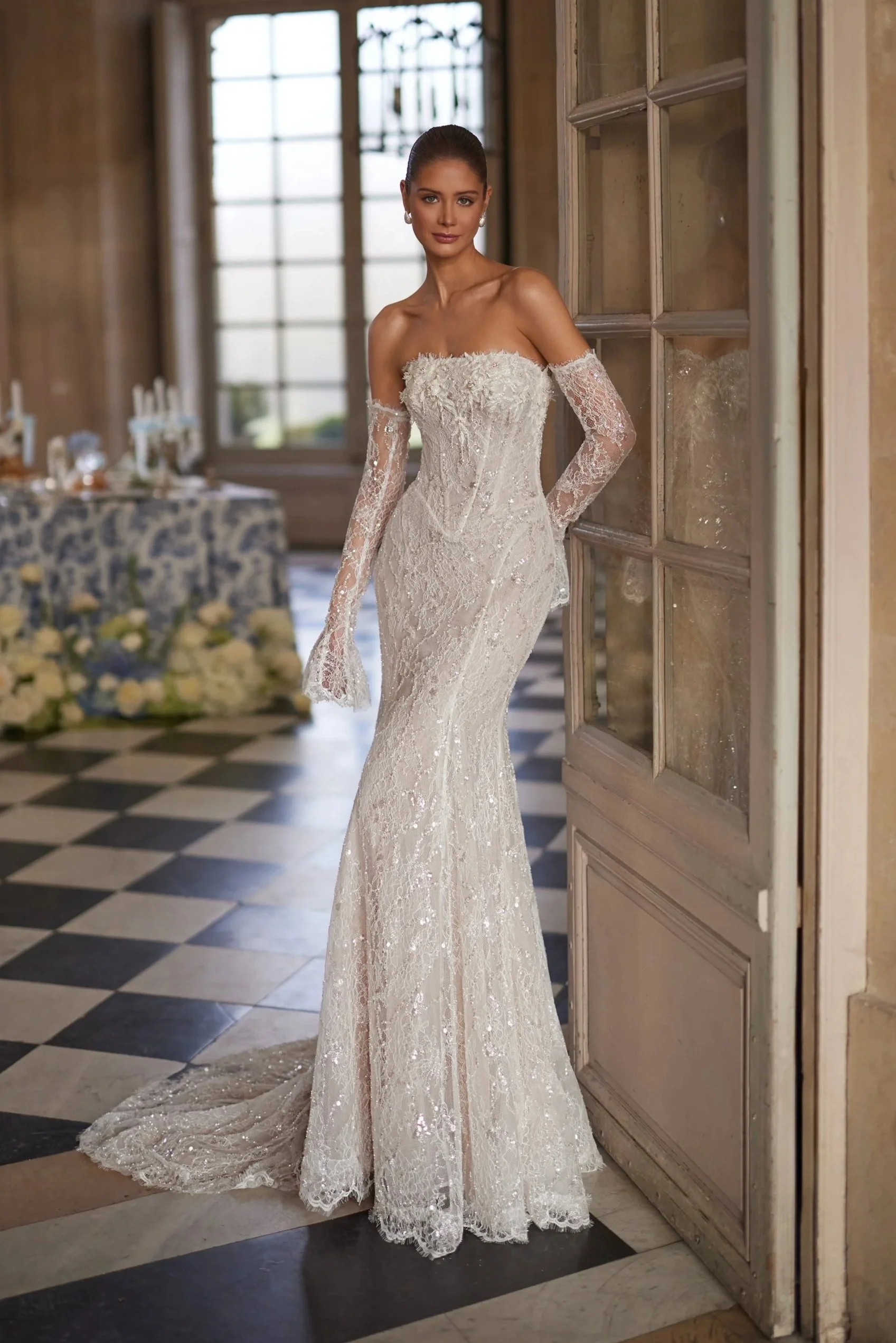 Frau in langem, weißem Hochzeitskleid mit Spitze, stehend in einem eleganten Raum mit großen Fenstern und Schwarz-Weiß-Fliesen.