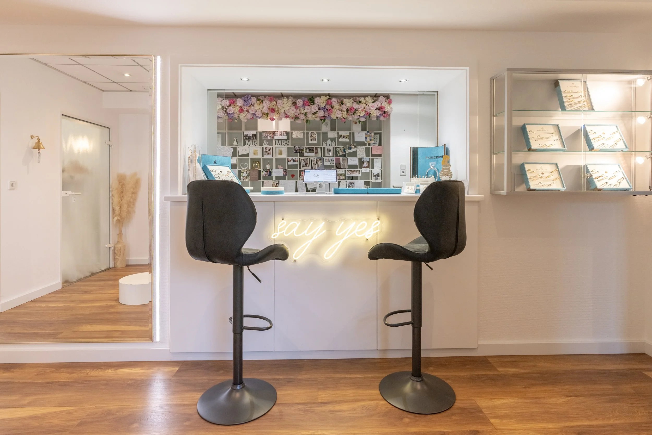 Ein Empfangstresen in einem hellen, modern eingerichteten Raum mit zwei schwarzen Barhockern, einer stilvollen Neon-Schrift, die 'say yes' sagt, und einer Wanddeko mit Fotos und Blumen. Links eine Tür, rechts eine Glasvitrine mit Schmuck.