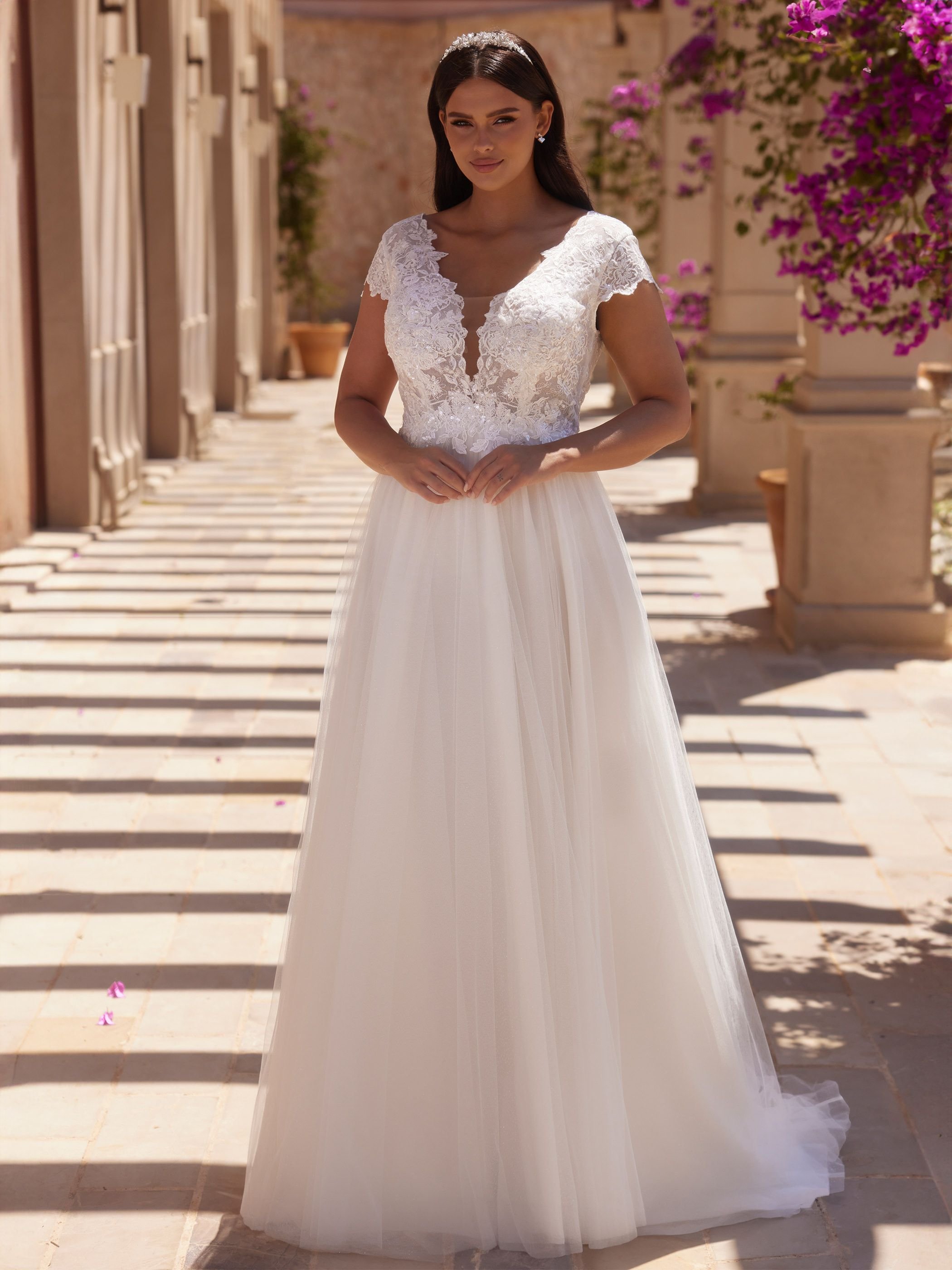 Frau in einem weißen, eleganten Brautkleid mit Spitzenoberteil steht in einem sonnendurchfluteten Gang mit Blumen im Hintergrund.