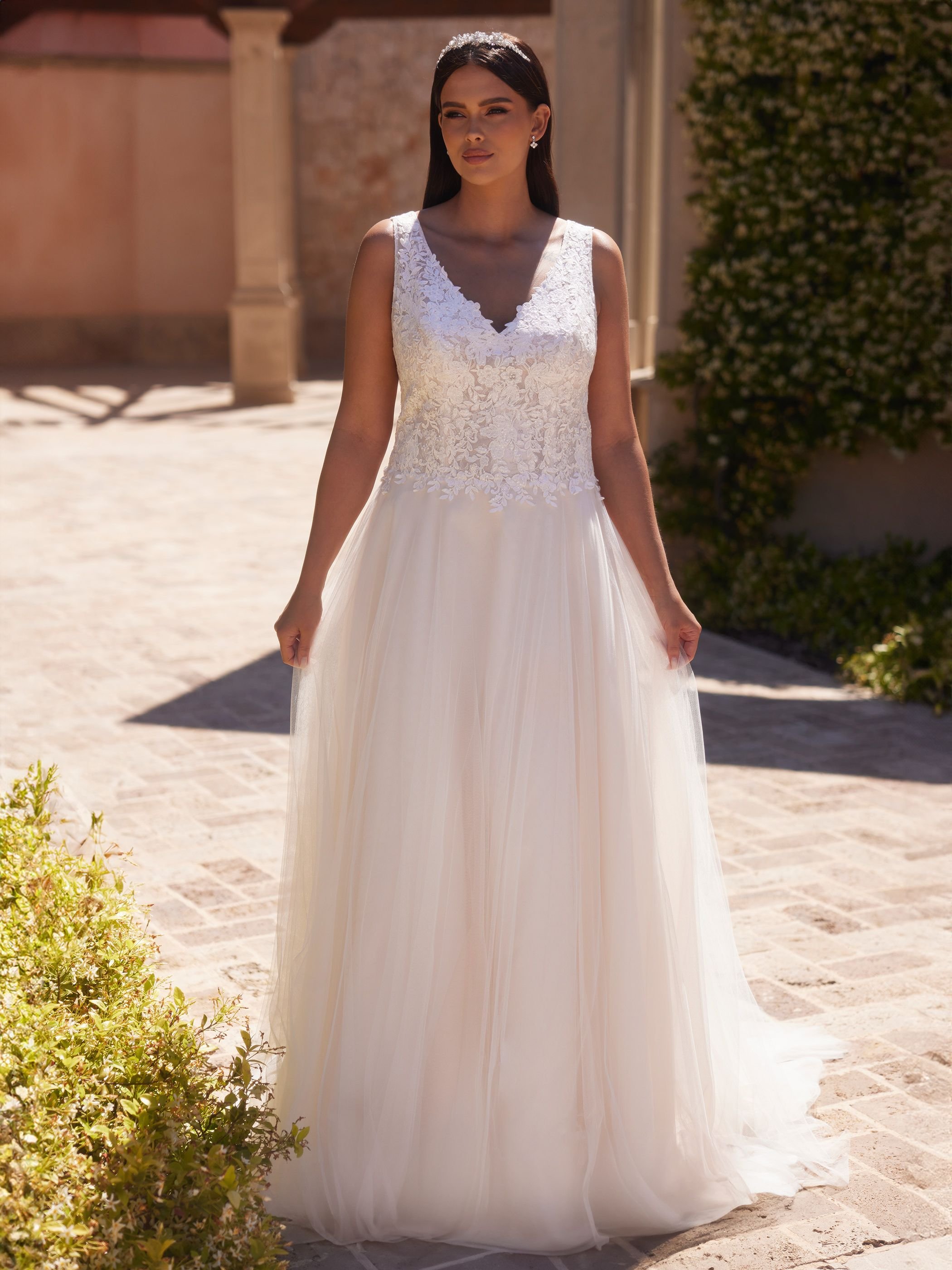 Eine Frau in einem Hochzeitskleid mit Spitze und Tüll steht draußen auf einem gepflasterten Weg, umgeben von Pflanzen.