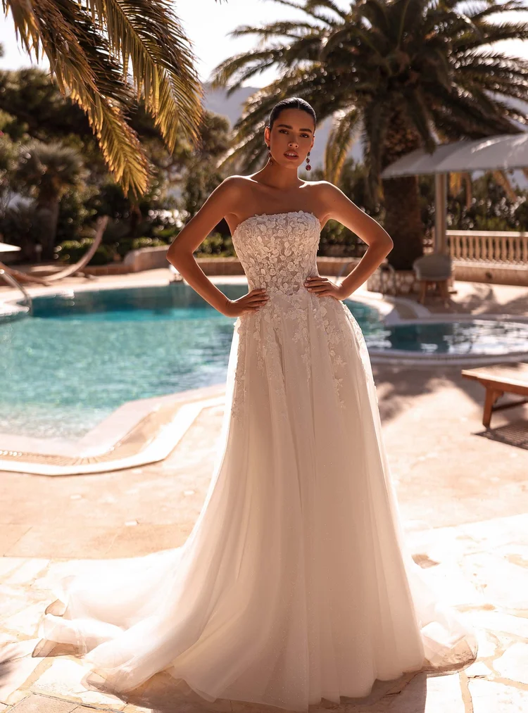 Frau im Brautkleid steht am Pool im Sonnenlicht, Palmen im Hintergrund, die Szenerie wirkt warm und luxuriös.