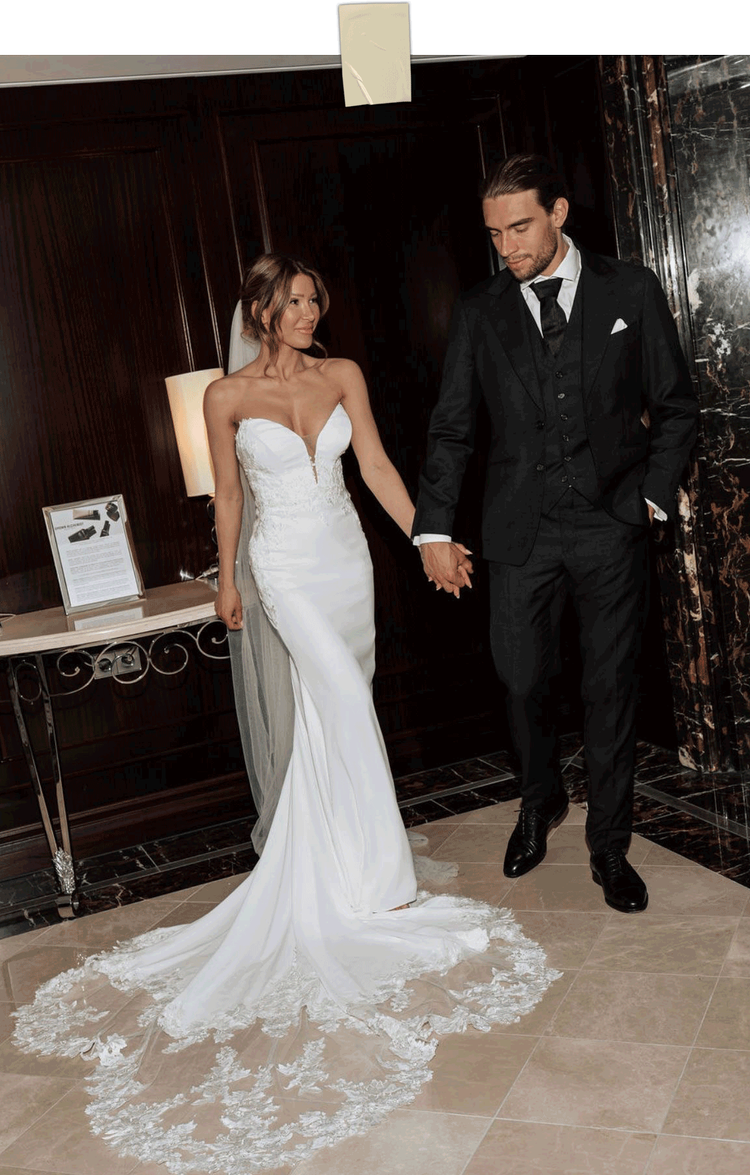 Ein Brautpaar hält Händchen und posiert in einem eleganten Raum, die Braut trägt ein langes weißes Hochzeitskleid mit Schleppe, der Bräutigam trägt einen schwarzen Anzug.