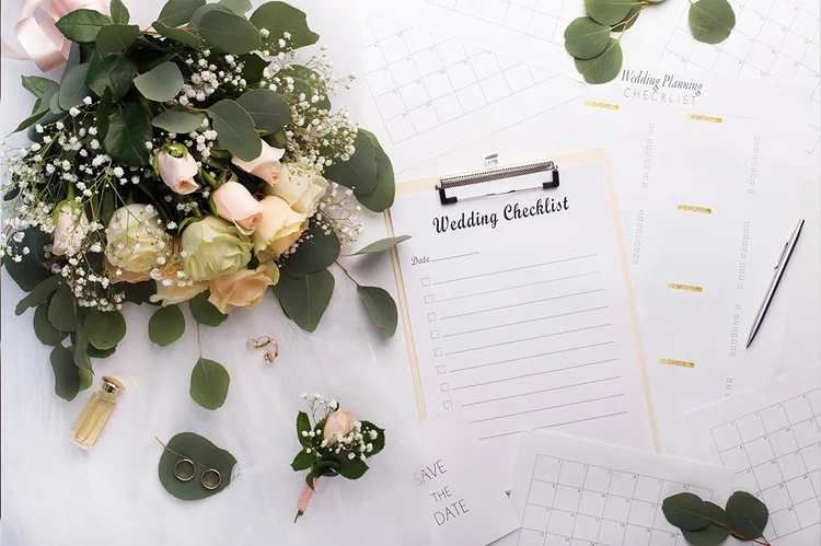 Blumenbouquet, Wedding-Checkliste, Notizblock, Stift, Schmuck, Parfümflasche und Eukalyptusblätter auf einem weißen Tisch, für Hochzeitsplanung.