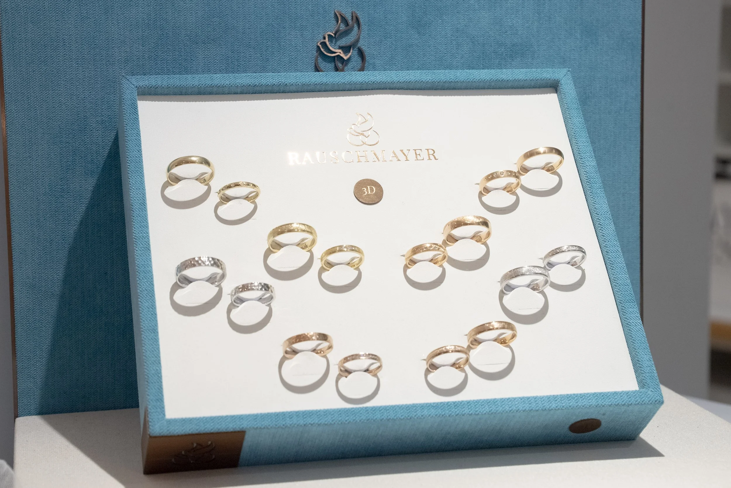 Blaue Schmuckschatulle mit goldenen und silbernen Ringen, mit der Aufschrift 'RAUSCHMAYER' und einem 3D-Siegel, die Ringe im Inneren sind sichtbar.