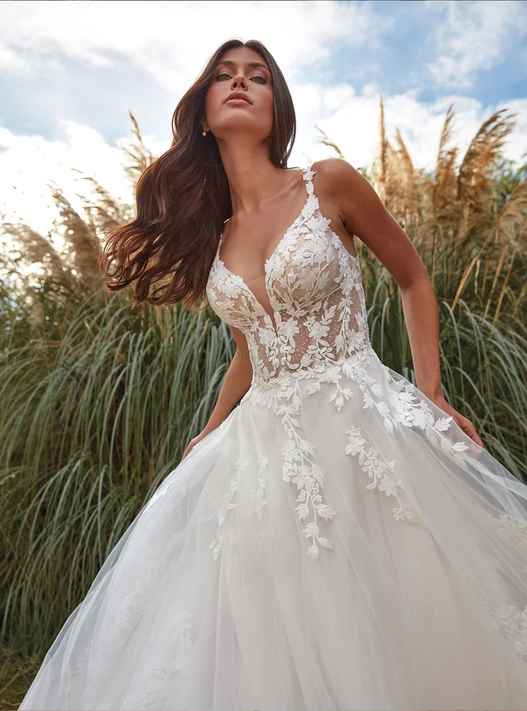 Eine Frau trägt ein weißes Hochzeitskleid mit floralen Spitzenmuster im Freien vor hohen Gräsern, unter einem bewölkten Himmel.