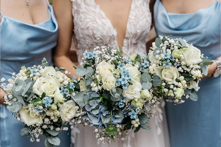 Drei Frauen halten Brautsträuße mit weißen Rosen, blauen Blumen, Eukalyptus und kleinen weißen Blüten bei einer Hochzeitsfeier.