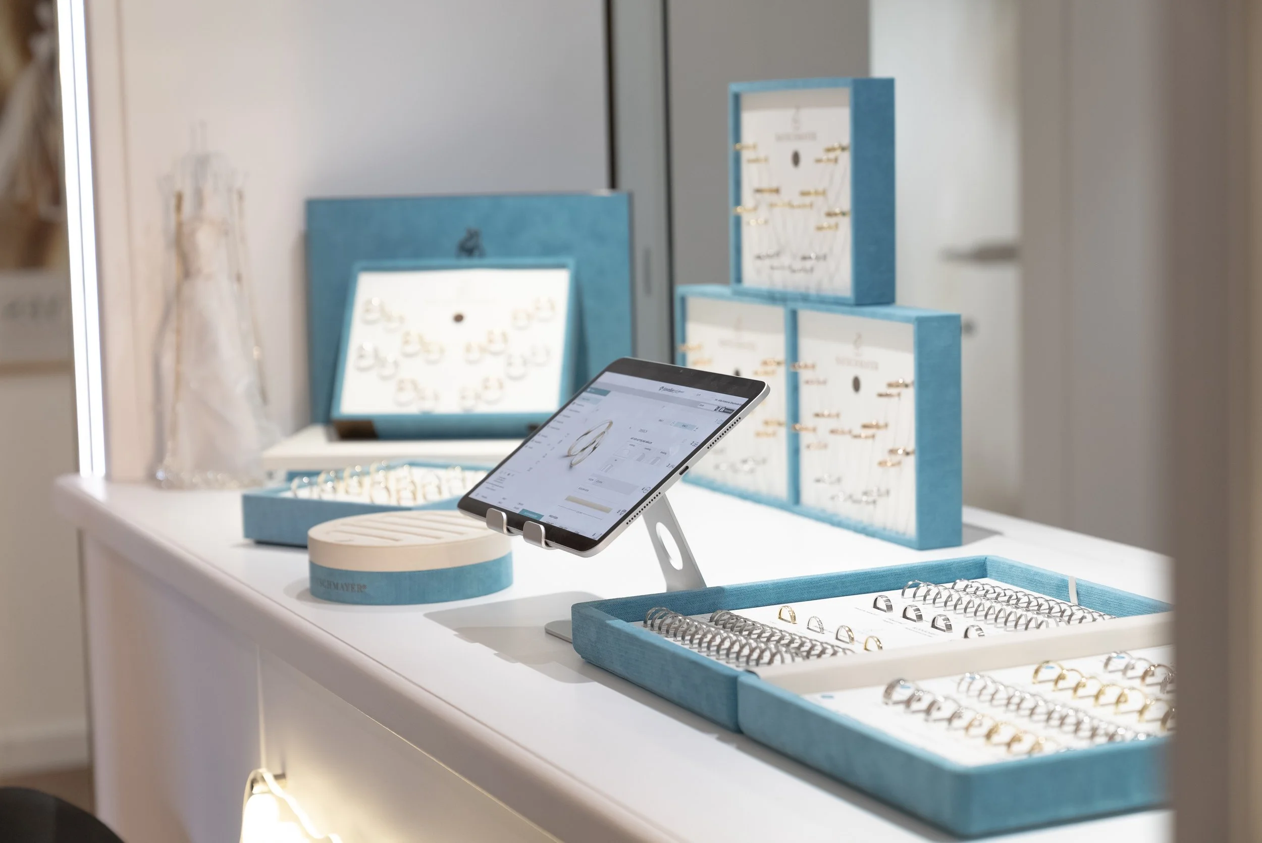 Ein Ausstellungsstand mit Schmuck in blauen Boxen, einem digitalen Tablet auf einem Ständer und weiteren Schmuckstücken in separaten Boxen, wahrscheinlich im Juweliergeschäft.