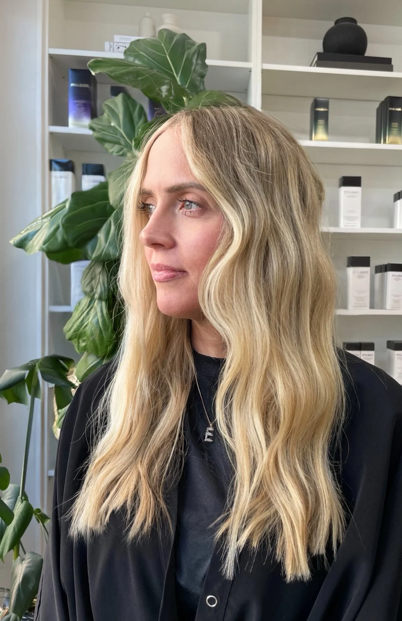 A woman with long wavy blonde hair looking to her left in a room with green plants and shelves of skincare products behind her.