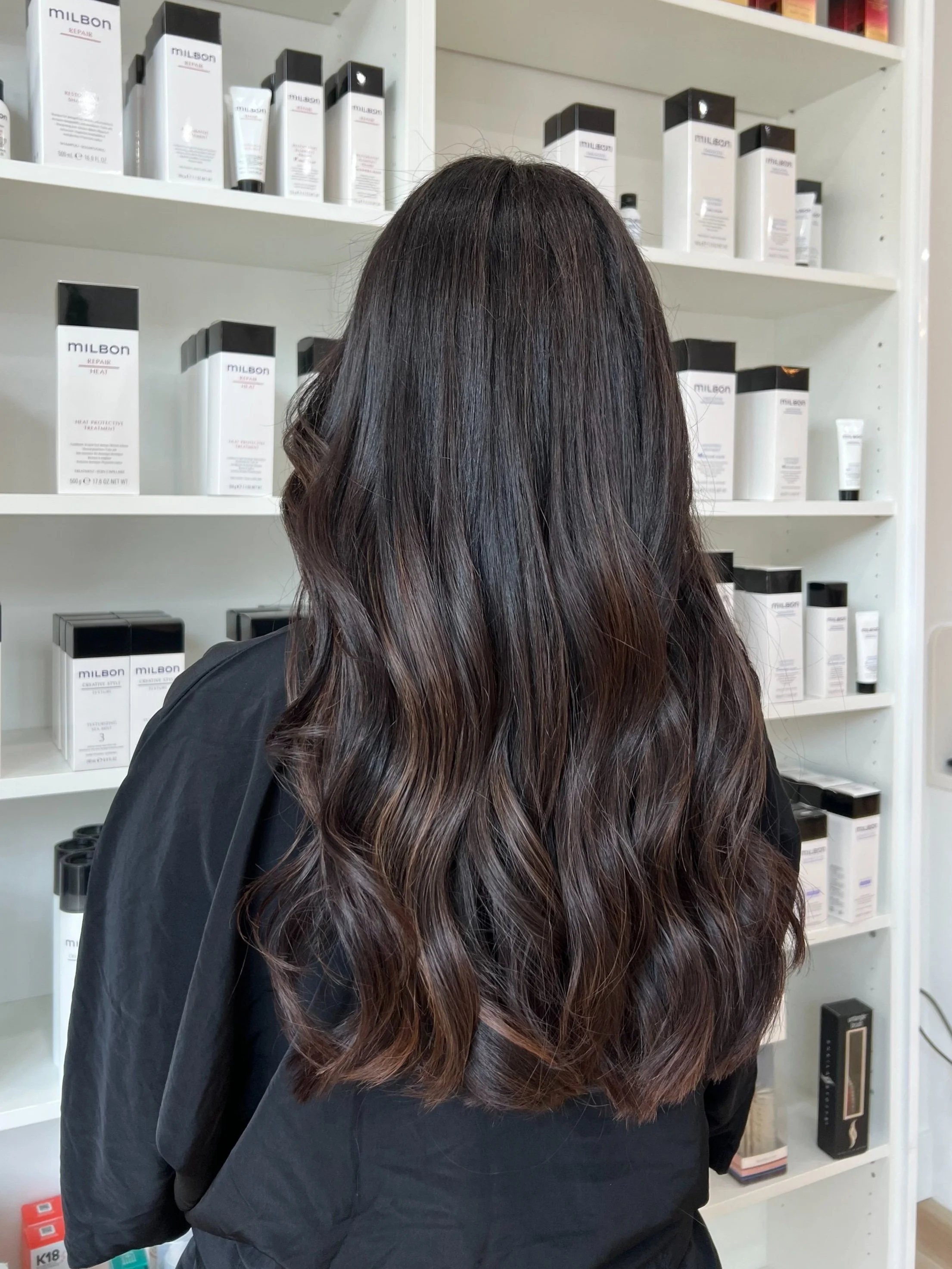Back of a woman with long, wavy brown hair standing in front of shelves filled with hair care products.