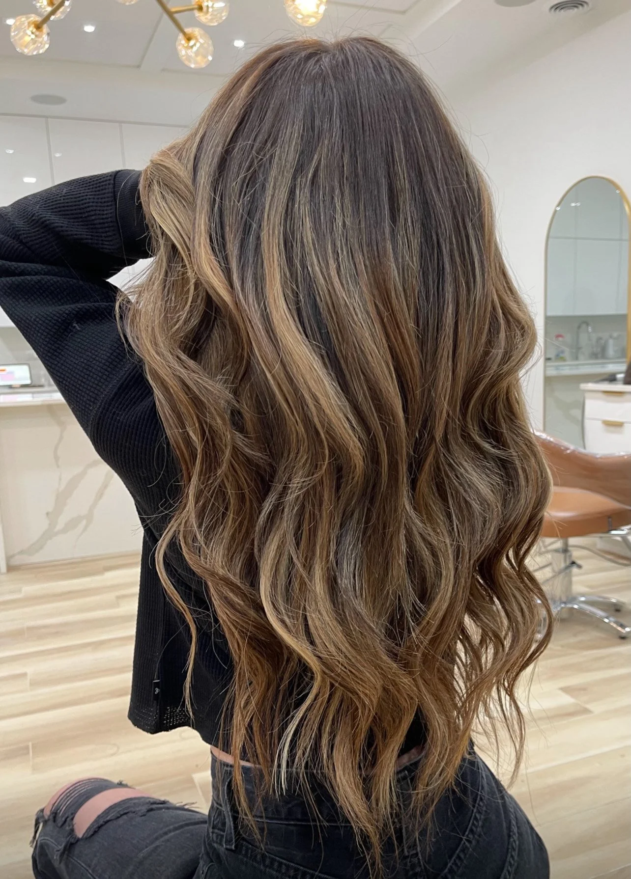 Woman with long wavy brown hair in salon, black outfit, warm lighting.