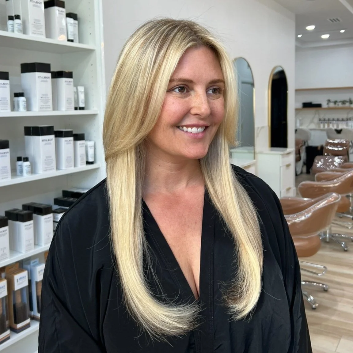 Blonde woman smiling in a salon with hair care products on shelf and salon chairs in the background.