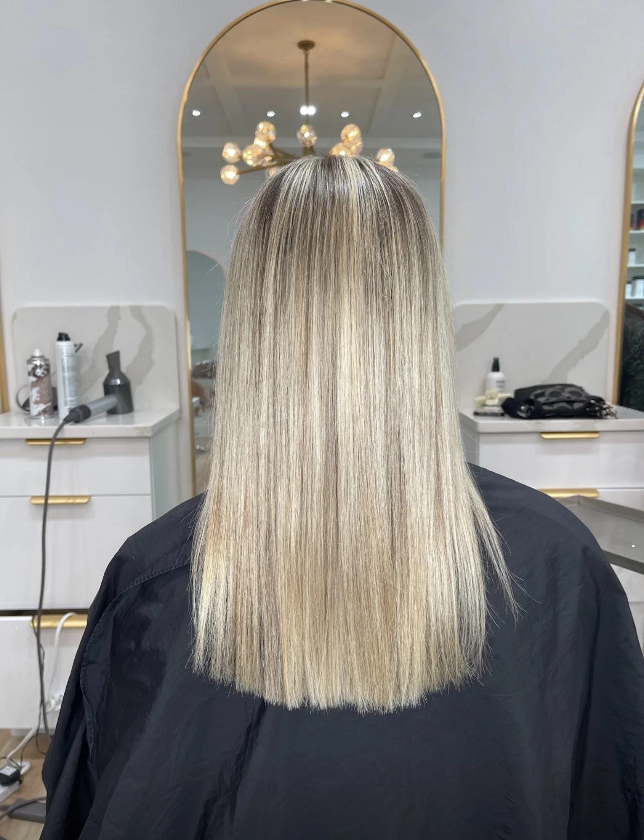 Back view of straight blonde hair in a salon with mirrors and styling tools.
