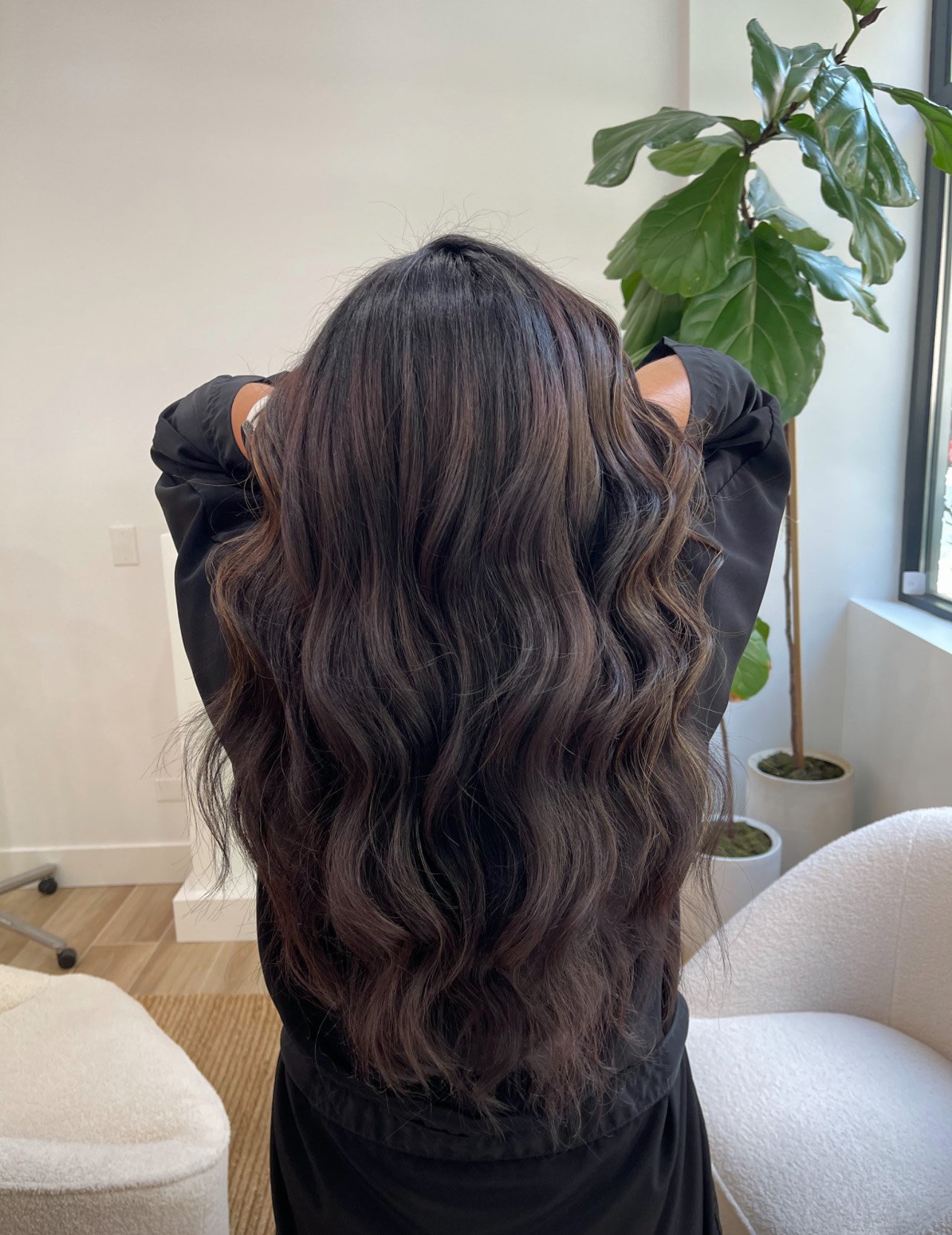 Person with long, wavy brown hair facing away, standing in a room with a green plant.