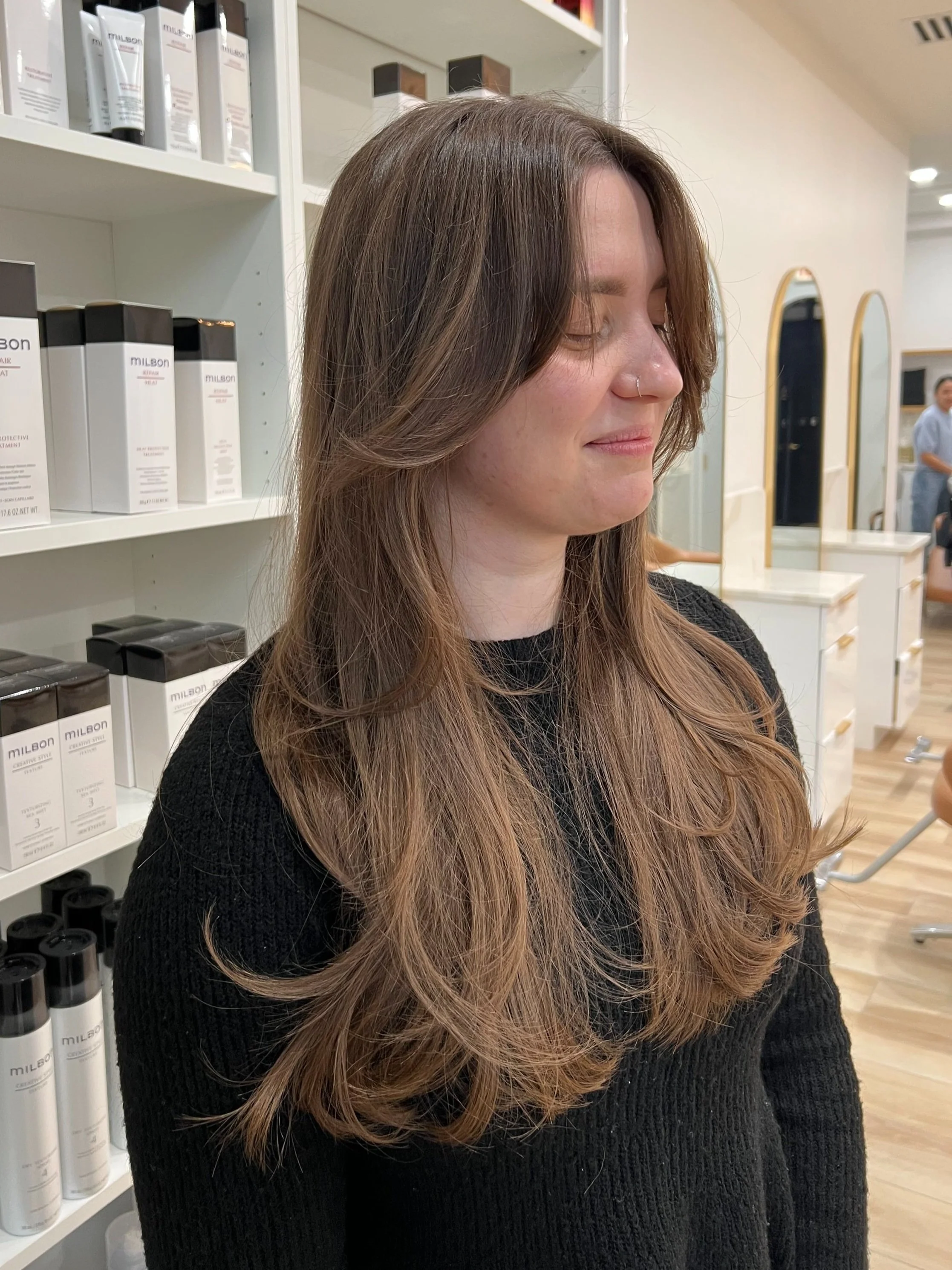 A young woman with long, wavy light brown hair and a nose ring, smiling with eyes closed in a hair salon with shelves of hair products in the background.