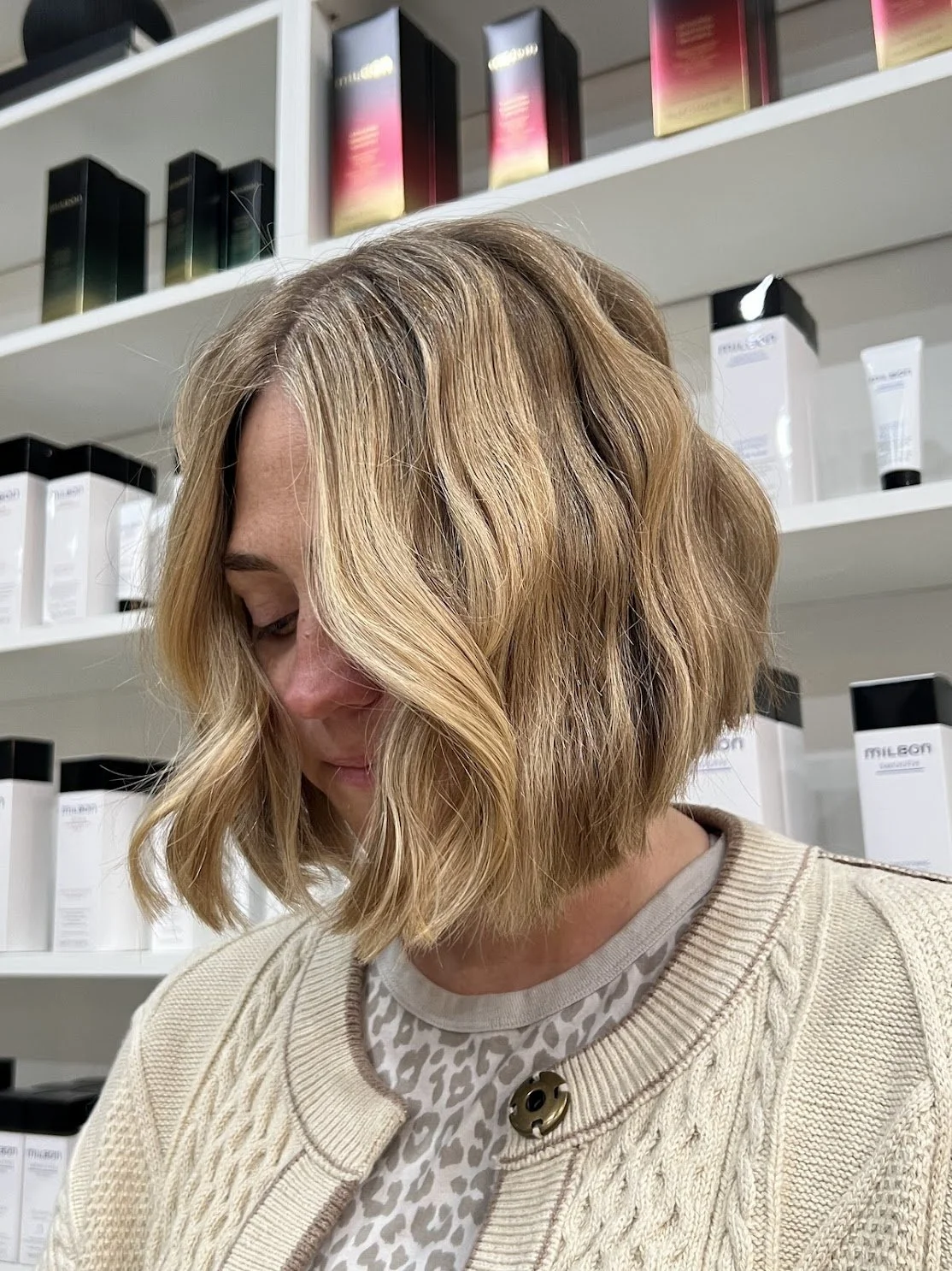 A woman with wavy blonde hair looking down, standing in front of shelves filled with hair or skincare products.
